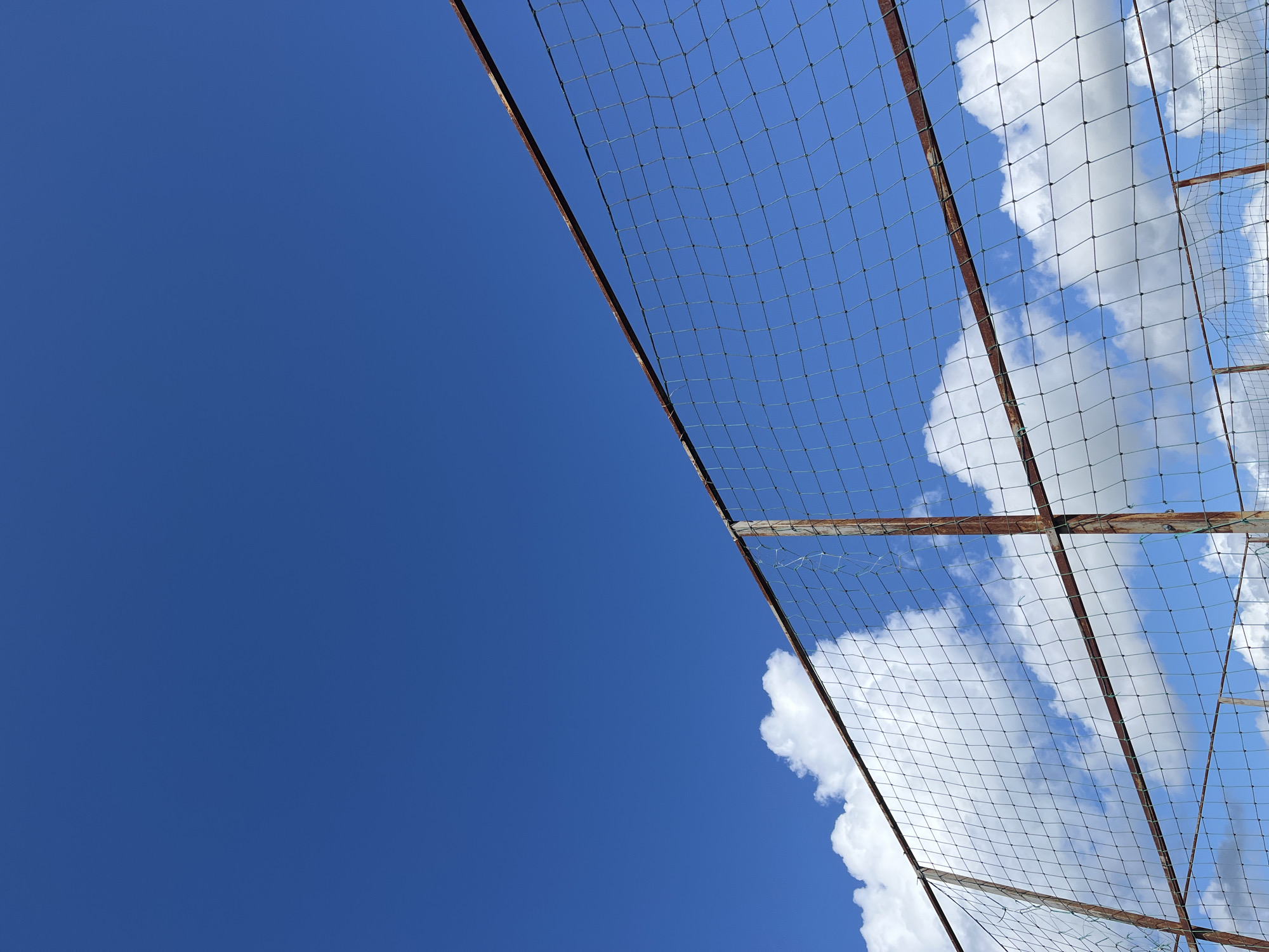 The image shows a section of a metal and mesh fence against a bright blue sky with scattered white clouds. The fence appears to be old and somewhat rusty, with visible metal posts and a netting material stretched across. The perspective is looking upwards, with the sky filling most of the frame. There are no people or discernible objects other than the fence and the sky. The scene appears to be outdoors. The location in Il'ino, Russia is not ascertainable from the image itself.