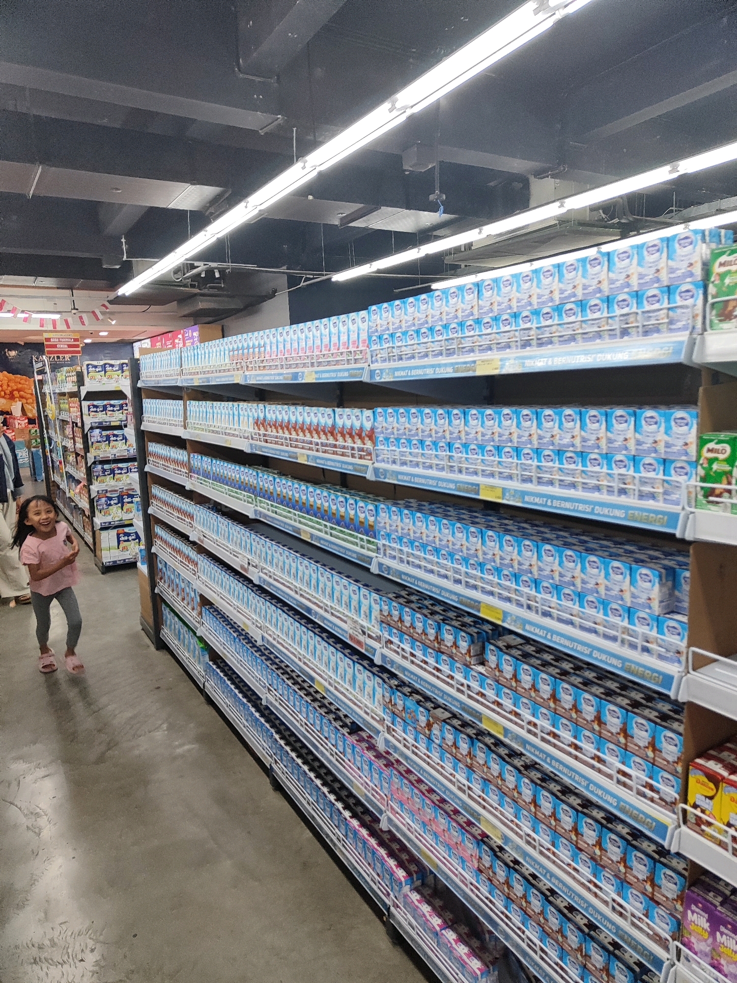 In a supermarket aisle in Sukowiryo, Indonesia, a young girl with dark hair, wearing a pink t-shirt and grey leggings, stands to the left of the frame, looking towards the camera with a wide smile. She is positioned on a polished concrete floor. To her right, shelves are densely packed with rows of blue and white cartons of milk, predominantly bearing the logo of SGM Eksplor. The cartons are arranged in multiple tiers, creating a visually repetitive pattern that extends down the aisle. Some shelves also contain brown cartons with red labels and green cartons with white labels, suggesting a variety of milk products are available. Overhead, fluorescent lights illuminate the scene, reflecting off the ceiling and the surfaces of the products. The overall impression is one of a well-stocked retail environment.