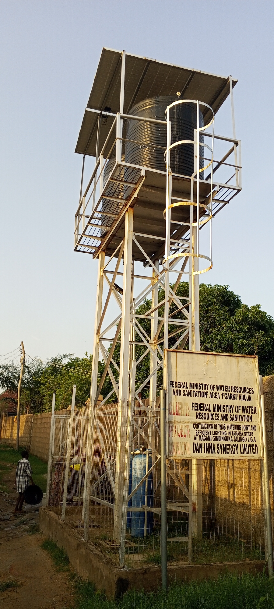 The image shows a water tower located in Jalingo, Nigeria. The tower is a metal structure supporting a large black water tank. It has a metal staircase leading up to a platform around the tank, and a canopy with solar panels overhead. A sign in front of the tower indicates that it is part of a project by the Federal Ministry of Water Resources and Sanitation, and that the contractor is Dan Inna Synergy Limited. The project involves the construction of seven watering points and efficient lighting at Magami Gindinkuka, Jalingo Local Government Area. In the foreground, a young boy in a checkered shirt is walking away from the camera, carrying a black bucket. A metal fence encloses the base of the tower, where a blue water tank or filter is visible. The environment appears to be a semi-urban or rural area, with a brick wall and trees in the background. The sky is clear and the lighting suggests it is late afternoon or early morning.