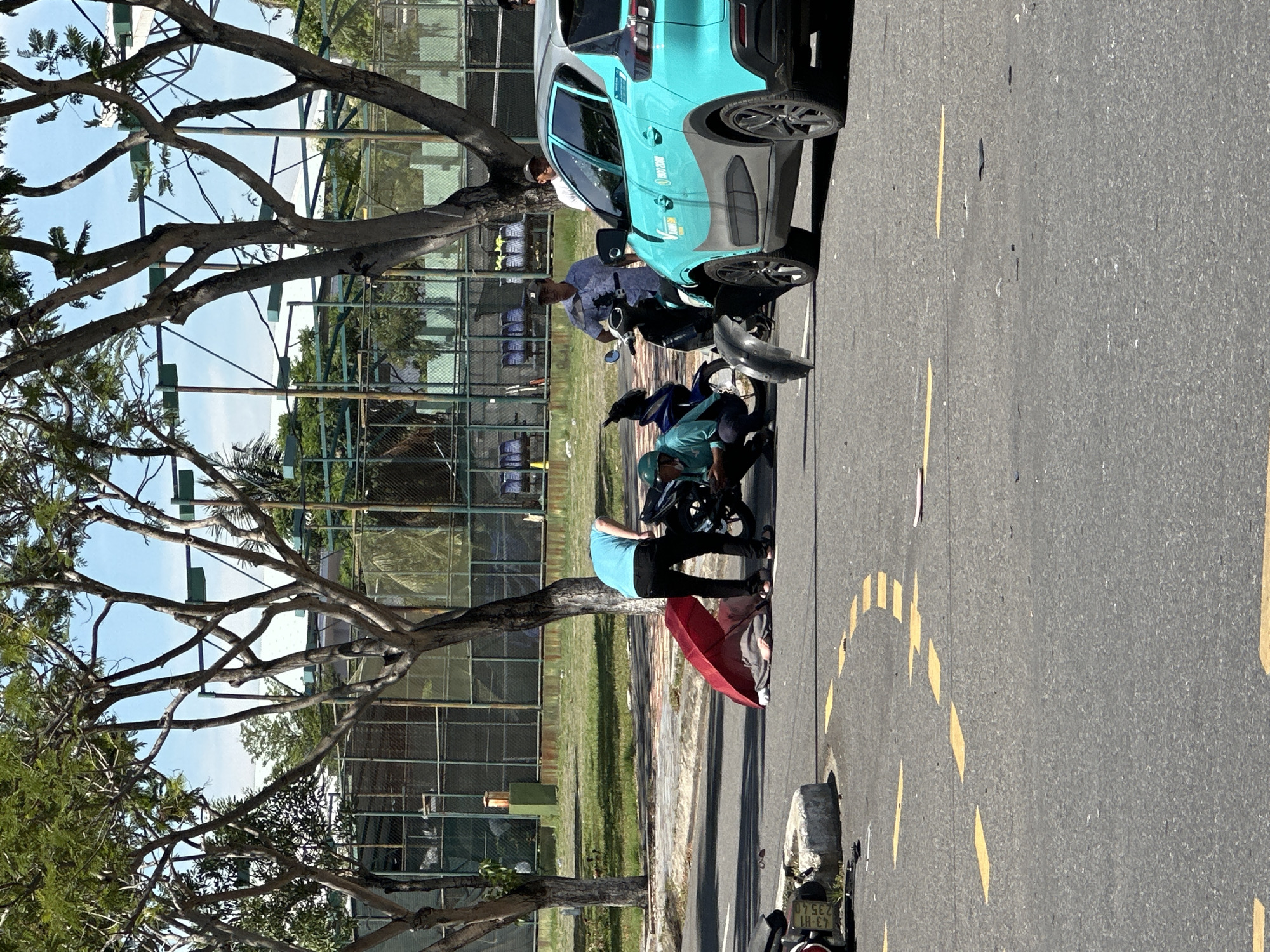 A scene of a traffic accident is depicted outdoors, likely in the city of Đà Nẵng, Vietnam, given the context. In the foreground, on a paved road with yellow lane markings, lies a person covered by a red umbrella, with another person in a teal shirt and dark pants kneeling beside them, seemingly attending to them. A blue and silver car, possibly a taxi, is parked on the right side of the road. A motorcycle, its front fender detached and lying on the road, is positioned between the car and the downed person. In the background, a chain-link fence surrounds a grassy area with trees. Several people are standing around, observing the scene. The weather appears sunny with a clear blue sky.