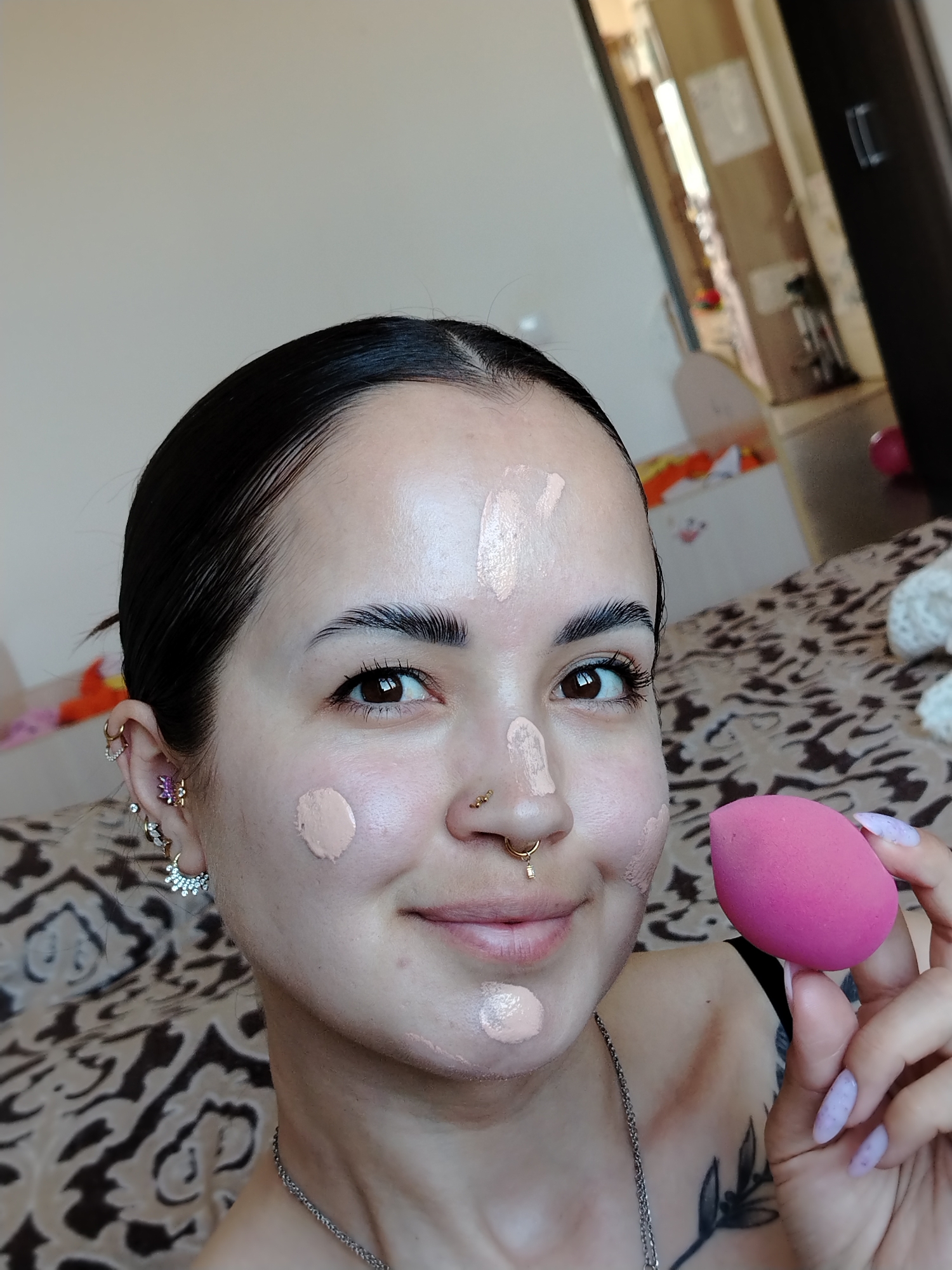 A fair-skinned person with dark hair pulled back is holding a pink makeup sponge in their right hand. Their face is covered in dots of light beige foundation. They have multiple ear piercings in their left ear and a gold nose ring. A silver chain necklace is visible around their neck. A tattoo is partially visible on their left shoulder. The background shows a patterned bedspread and a glimpse into another room. The city name is not discernable from the image.