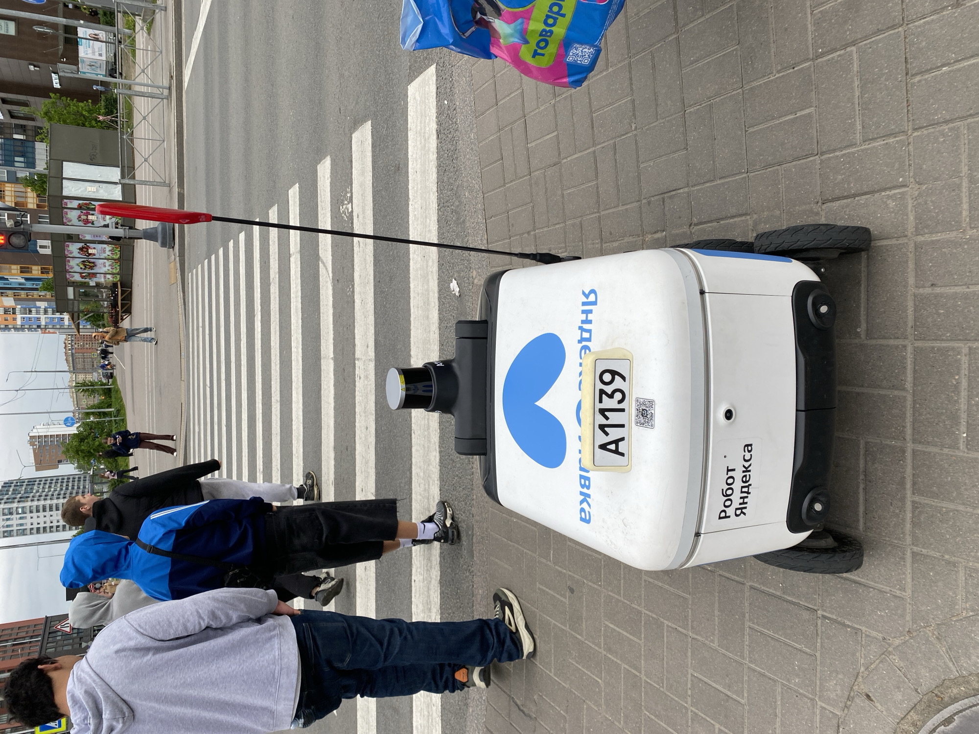 Here is a detailed description of the image:
**Location:** The image is set in Saint Petersburg, Russia, identifiable by the Cyrillic text on the Yandex delivery robot.
**People:** A group of people are crossing a pedestrian zebra crossing. Individuals are wearing casual clothes.
**Subject:** The primary subject is a white Yandex delivery robot. It has a blue heart logo and an identification number "A1139". The robot is equipped with wheels, a sensor, and a red flag.
**Objects:** Other objects include a shopping bag above the robot, a crosswalk, buildings in the background.
**Actions:** People are walking across the crosswalk, and the robot is stationary at the curb.
**Interactions:** The robot is positioned near pedestrians on the crosswalk, possibly preparing to cross or having just completed crossing.
**Environment:** The scene is outdoors, during daylight. The ground is paved. The background includes buildings.