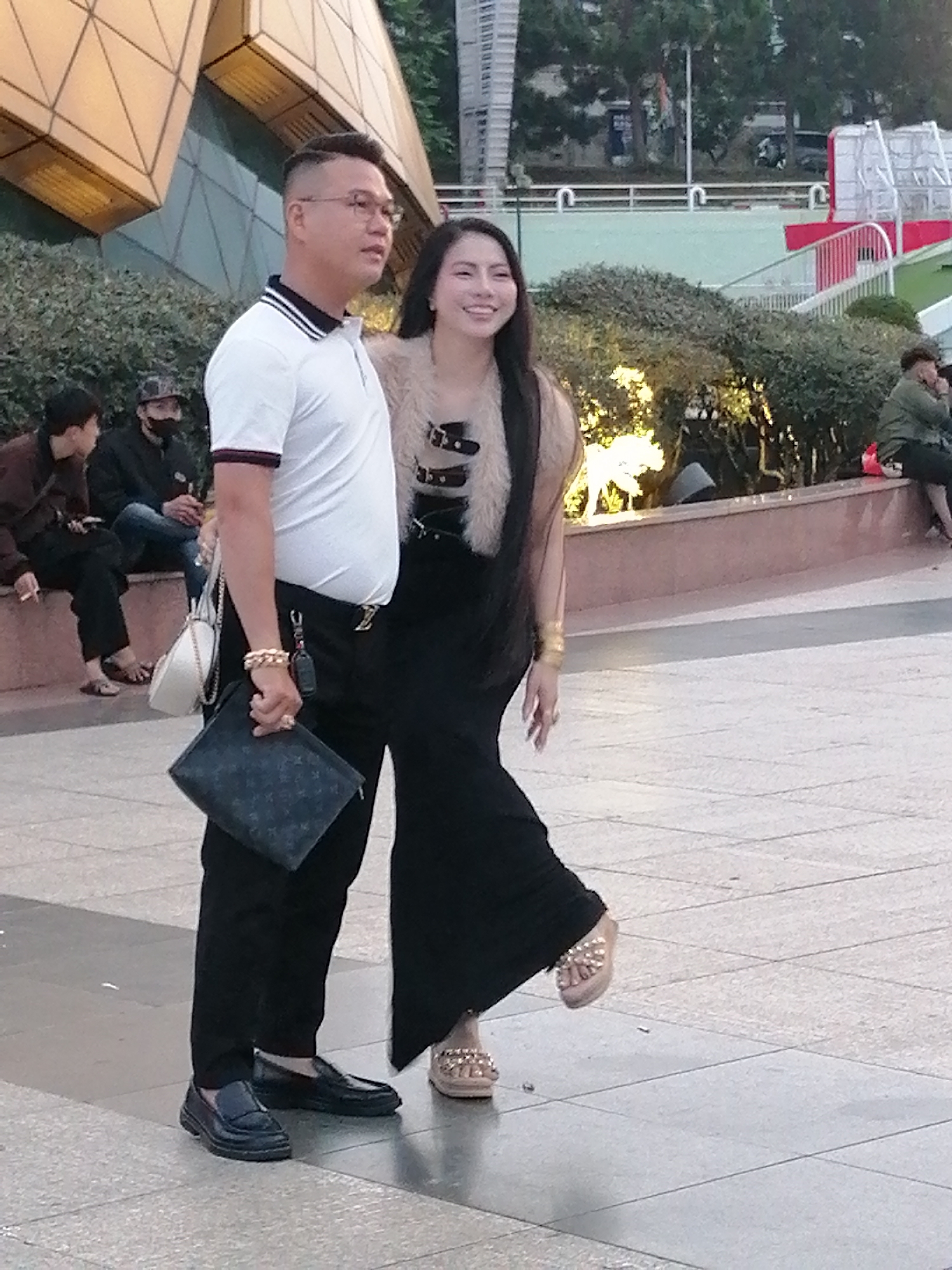 The image captures a man and a woman standing in what appears to be a public plaza in Da Lat, Vietnam, with a distinctive modern building with a geometric facade visible in the background. The man, wearing glasses, a white polo shirt with striped collar and cuffs, black pants, and black loafers, is holding a dark patterned clutch bag and a small white purse. He has a thick gold bracelet on his left wrist. The woman, with long dark hair, is wearing a black wide-leg jumpsuit, a light-colored fur-trimmed vest with buckles, and gold embellished sandals. She is smiling and looking towards the viewer. In the background, several other people are seated on a tiered stone wall, and some are walking. The plaza is paved with large square tiles. The environment suggests an urban setting, possibly a tourist area, given the architectural style and the presence of multiple individuals.