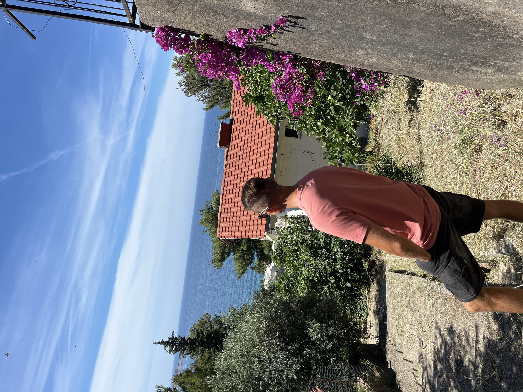 A male stands with his back to the camera, facing the Adriatic Sea in Šušanj, Montenegro. He wears a short-sleeved coral-colored t-shirt and black shorts. The man stands on a sun-drenched, dry grassy slope with scattered stones and foliage. In the mid-ground, a white house with a red-tiled roof is visible, surrounded by lush green trees and vibrant pink bougainvillea climbing a stone wall. The clear blue sea stretches to the horizon, meeting a bright blue sky dotted with wispy white clouds. Several birds are visible in the sky. The scene is set on a sunny day.