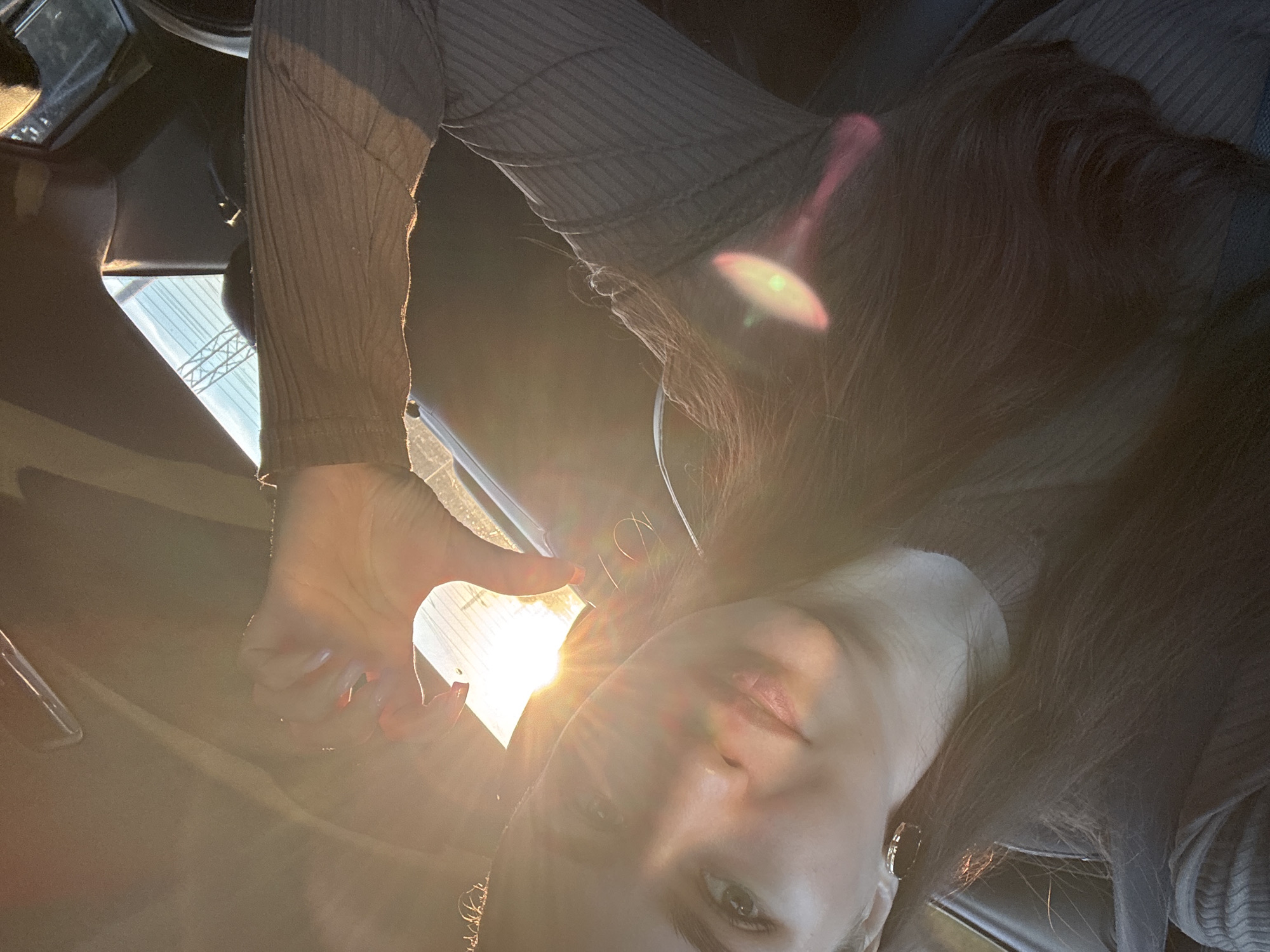 This image is a selfie-style portrait of a young woman taken inside a vehicle. The woman, with long, dark hair and wearing a dark, ribbed long-sleeved top, is seen from the chest up, with her head tilted as she looks towards the camera. Her face features light-colored eyes and full lips, softly illuminated by the golden light. An earring is visible on her right ear.
Her left hand, adorned with red nail polish, is raised to form a heart shape with her thumb and index finger, suggesting a warm or affectionate sentiment.
A strong, warm light source, likely the sun, streams in from the right side of the frame, creating prominent lens flares and backlighting her. This intense, low-angle sunlight indicates either sunrise or sunset, suggesting clear and sunny weather conditions. The setting appears to be the interior of a car or bus, with dark upholstery visible. Through a window in the background, a light blue sky and a faint outline of a metallic structure, possibly part of a bridge or industrial architecture, can be seen. The overall atmosphere is intimate and positive, illuminated by the bright, natural light. No text is visible in the image. The location context for this scene is Chaikovsky, Russia.