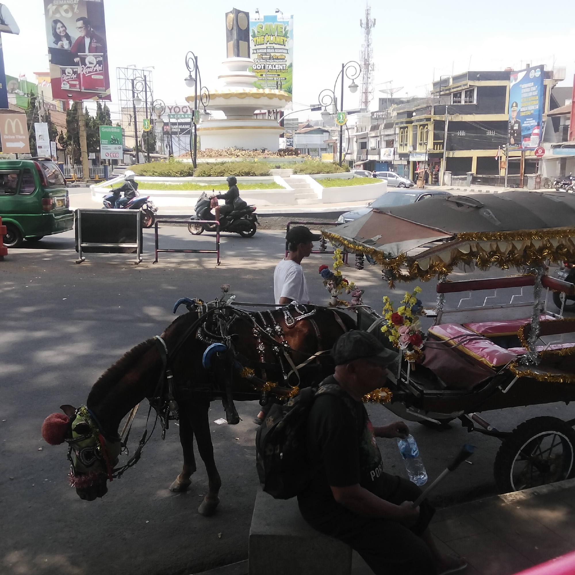 In Sukabumi, Indonesia, on a sunny day, a horse-drawn carriage is parked on an asphalt road. The carriage is adorned with gold tassels and colorful flowers, with red and pink cushions visible inside. A dark-colored horse with green and red decorations on its head is harnessed to the carriage. Two men are associated with the carriage; one, wearing a black cap and a black t-shirt with "PORT" visible on the back, sits on a concrete block, holding a bottle of water and a stick. Another man, wearing a white t-shirt and a black cap, stands near the carriage, looking to his right. In the background, a traffic circle features a white, tiered monument topped with a golden structure. Surrounding the traffic circle are various buildings, including those with black and yellow facades, and a tall radio tower. Billboards are also visible, including one with an image of a couple and text in Indonesian, and another promoting "SAVE THE PLANET." Several motorcycles and a green vehicle are present on the road, indicating a typical street scene in the city.