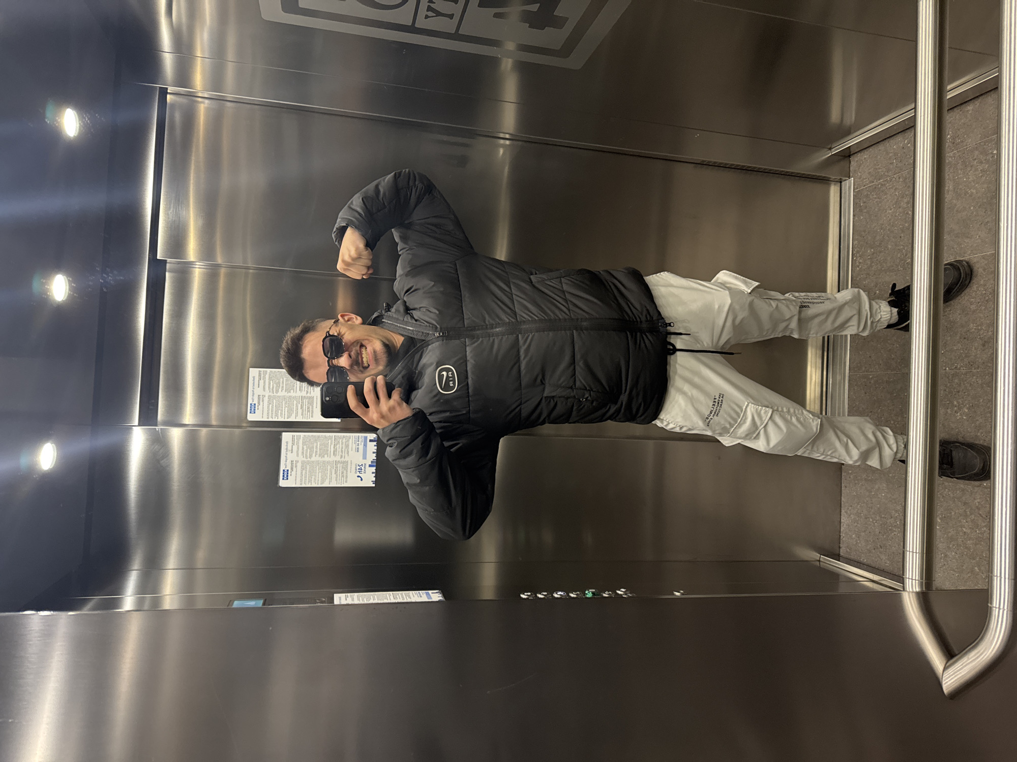 A man in sunglasses and a black puffer jacket poses in an elevator. He is flexing his bicep while holding a phone, apparently taking a selfie. He is wearing white pants and black shoes. The elevator has stainless steel walls, with informational signs posted on one wall and elevator buttons visible. The location appears to be in Sofia, Bulgaria, based on the Cyrillic alphabet visible on one of the signs.