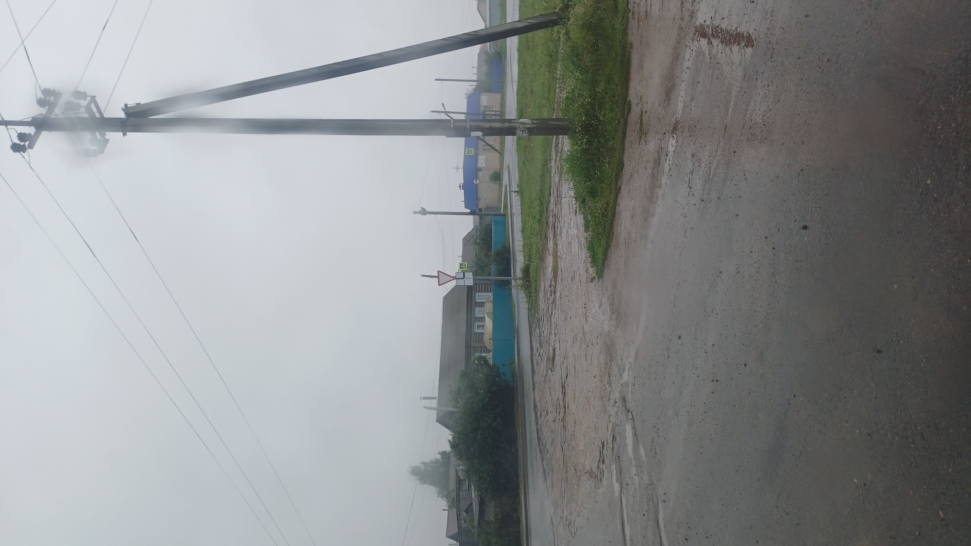 The image shows a street scene in the village of Kiyasovo, Russia, on a cloudy, overcast day, possibly with rain, as indicated by the wet pavement and reflections. In the foreground, a wet asphalt road curves to the left. To the right of the road is a patch of green grass and a power pole with multiple wires. In the middle ground, there is a cluster of wooden houses with dark roofs and a blue fence. A yield sign and a pedestrian crossing sign are visible near the road. Further in the background, more houses and trees are discernible through the hazy atmosphere. The overall impression is of a quiet, rural setting experiencing inclement weather.