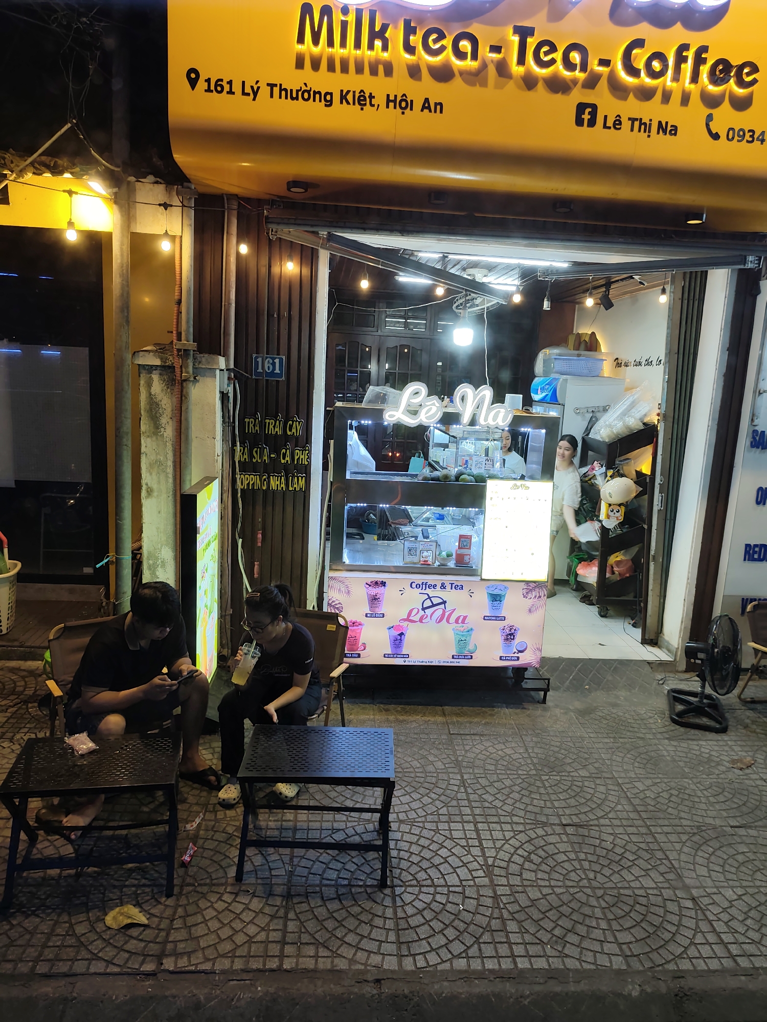 In this nighttime street-level shot of a storefront in Unknown, Unknown, a yellow awning with the words "Milk tea-Tea-Coffee" in illuminated lettering extends across the top of the frame. Beneath the awning, a doorway leads into a brightly lit interior. In front of the storefront, two individuals are seated in portable chairs at small tables. The person on the left, a man with dark hair, is looking down at his phone, while the person on the right, a woman with glasses and dark hair pulled back, is holding a drink with a straw and appears to be sipping it. A refrigerated display case in the center of the frame showcases various beverages with illustrated cups. A person can be seen working behind the counter inside. The ground is paved with patterned concrete tiles. A black oscillating fan is positioned to the right of the storefront.