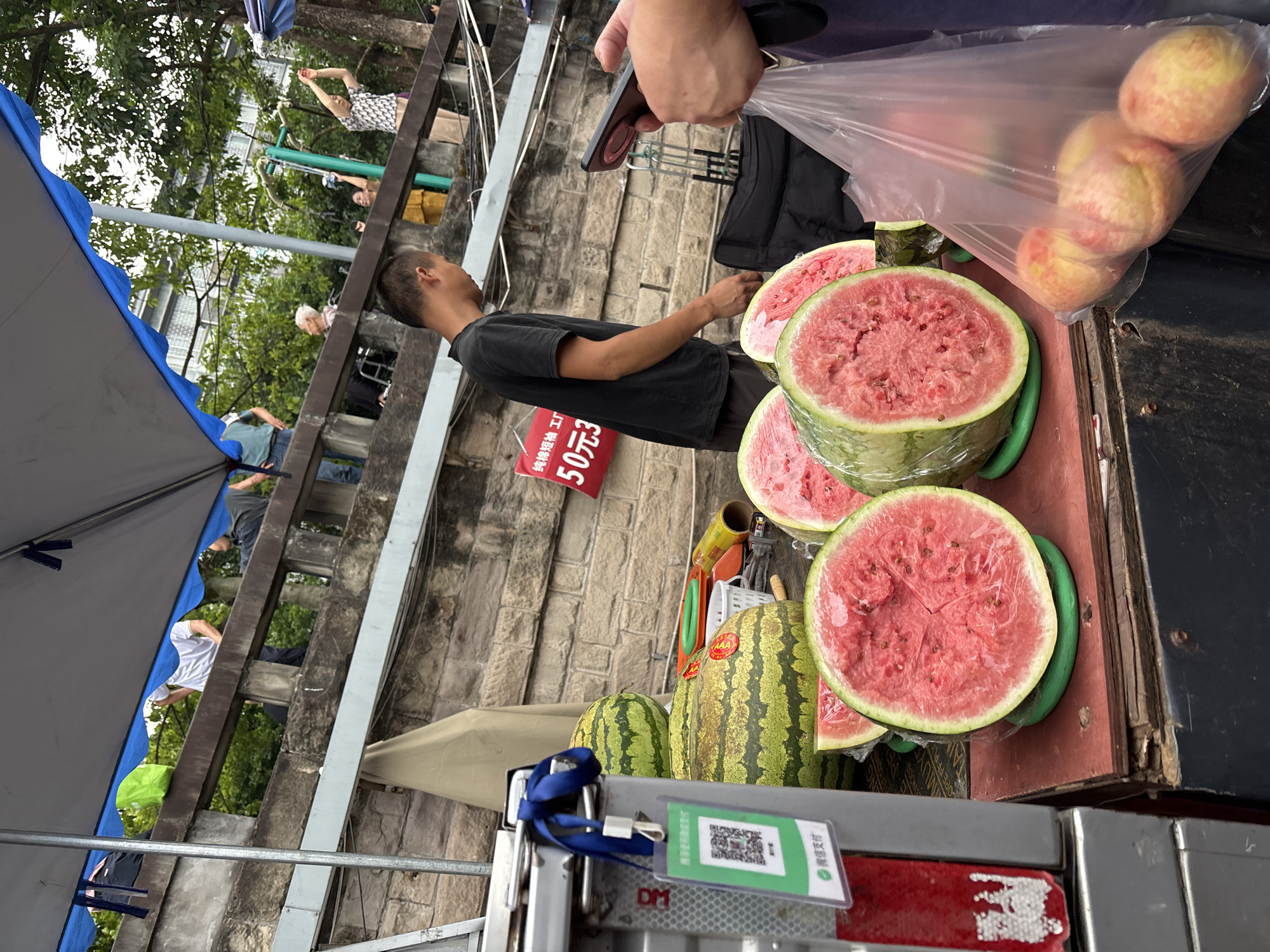 A street market scene in Jiangjin, China, features a vendor with a selection of watermelons and other produce. Several whole watermelons, some halved and wrapped in plastic, are displayed on a wooden surface. A man in a black t-shirt stands behind the produce, facing away from the camera. To the right, a person holds a mobile phone and a plastic bag containing several peaches. In the background, a stone wall is visible, with people on a higher level observing the market. A red sign with Chinese characters and the number "50" is mounted on the wall. The overall environment suggests an outdoor market setting with a variety of goods for sale.