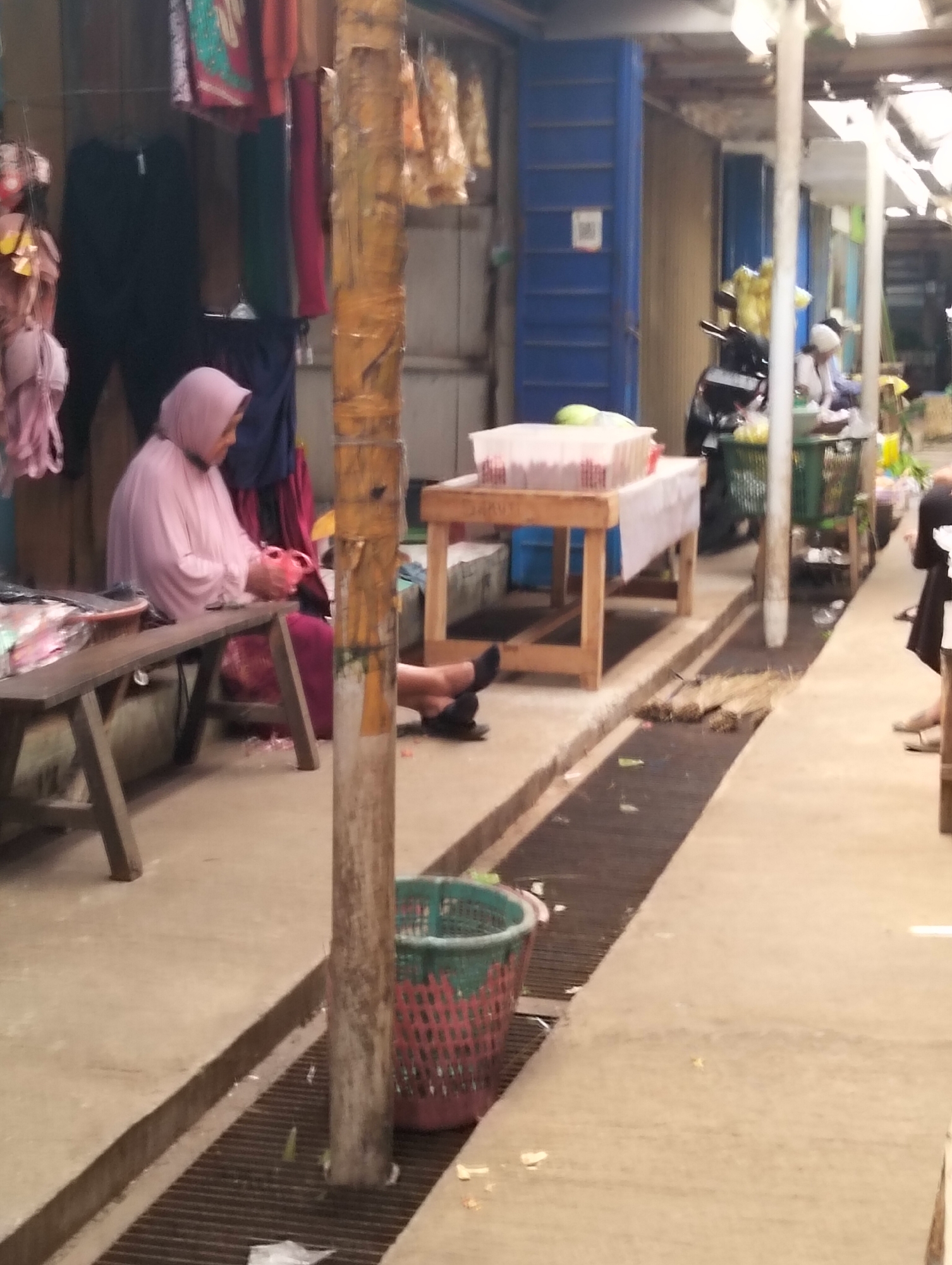 The image depicts a market scene in Subah, Indonesia. A woman wearing a pink hijab and pink top is seated on a wooden bench, appearing to be organizing or handling some items. In front of her, on the bench, is a plastic container and some loose items. To her left, a wooden table with a white cloth is set up, displaying another clear plastic container filled with what appears to be food items. Behind the woman, several garments are hanging, including black pants and colorful fabrics. A wooden pole with a rough texture stands prominently in the foreground, partially obscuring the scene. In the background, there are market stalls with blue shutters, a motorcycle, and various baskets. The ground is concrete, with a drainage grate running along the pathway. The overall impression is of a busy, everyday market environment.