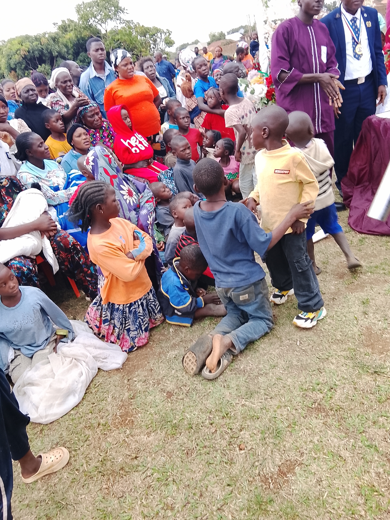 A gathering of people, including children, is taking place outdoors in Kuru, Nigeria. The individuals are diverse in age and attire, with some seated and others standing. Several children are playing or interacting near the front of the group. The setting appears to be a grassy area, possibly a park or open space, with trees visible in the background. Clothing styles are varied, with traditional African fabrics mixed with modern garments. Two men stand slightly apart from the larger group.