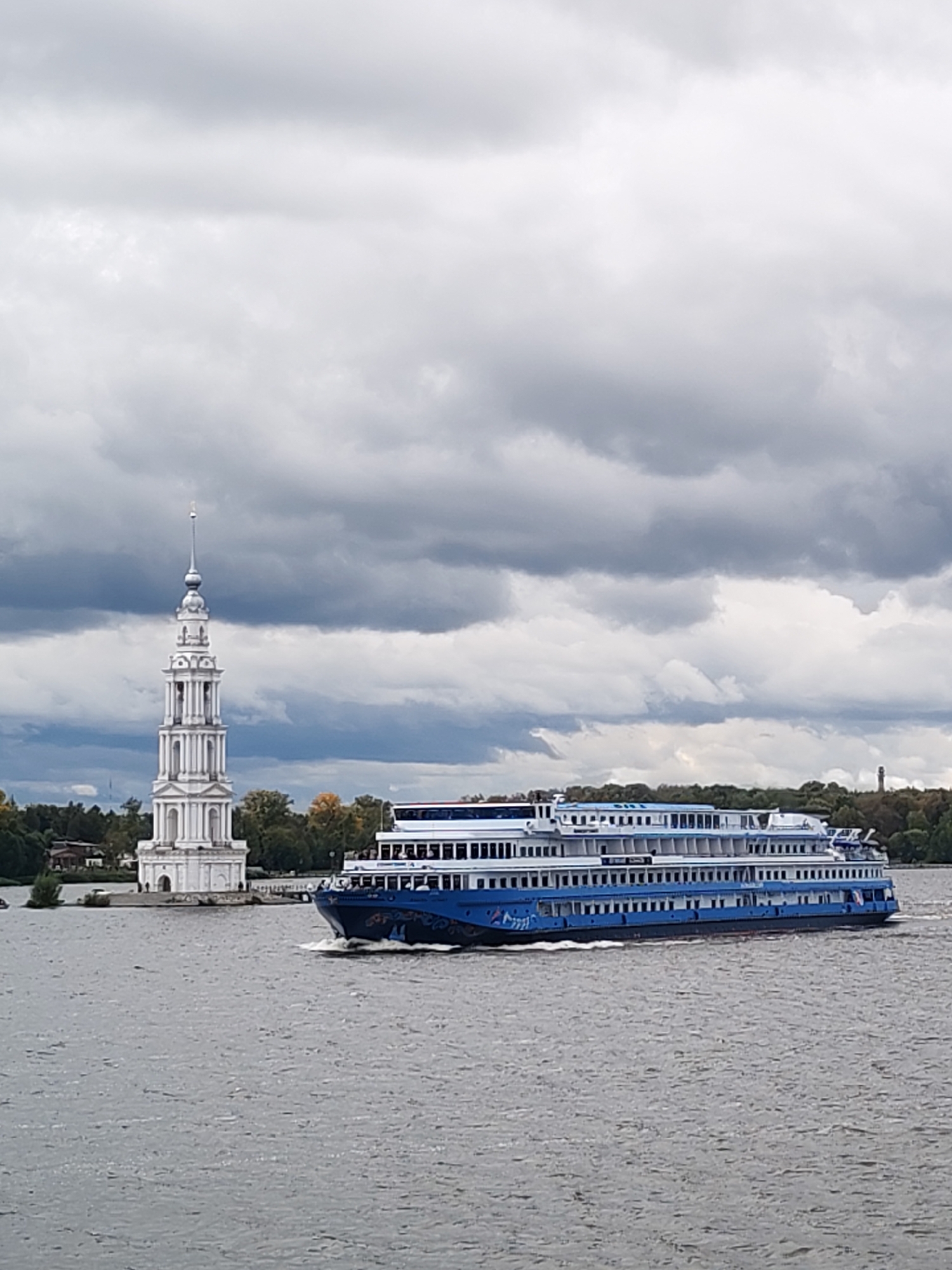 The image shows a wide river with a large, multi-deck passenger ship sailing on it. The ship is predominantly white with blue accents and is moving from right to left. In the background, on the left side of the image, is a prominent white bell tower with a spire, characteristic of the Kalyazin bell tower in Kalyazin, Russia. The bell tower stands on a small island or peninsula, with some trees and buildings visible behind it. The sky is overcast with dramatic, dark clouds. The water is choppy, indicating movement on the river. The overall scene depicts a calm but potentially dramatic day on the Volga River near Kalyazin, with the iconic bell tower and a modern passenger vessel as the main subjects.