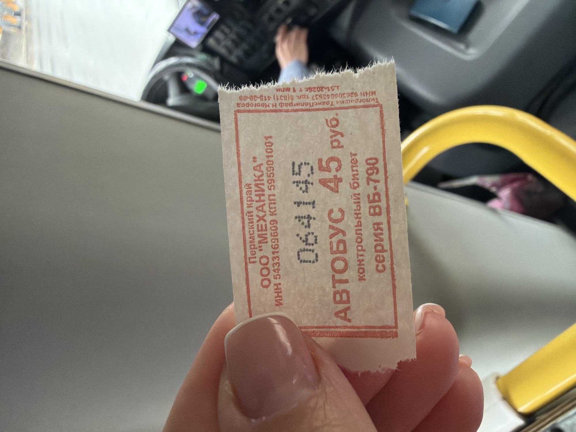 A close-up shot shows a person's hand holding a used bus ticket inside a vehicle, likely a bus. The bus interior is visible in the blurred background, featuring a yellow grab rail and a glimpse of the driver's area with controls and a person's hand on the steering wheel or dashboard. The lighting suggests an indoor setting, possibly during the day or evening.
The white paper bus ticket is slightly tattered at the top edge and features printed text in red. Key details on the ticket include:
* "Пермский край" (Perm Krai), indicating the region.
* "ООО "МЕХАНИКА"" (LLC "MEKHANIKA") with associated tax identification numbers (ИНН and КПП).
* A ticket number "064145".
* "АВТОБУС 45 руб." (BUS 45 rub.), specifying the fare and mode of transport.
* "контрольный билет" (control ticket) and "серия ВБ-790" (series VB-790).
* The printer's information: "Типография Транс Полиграф Н. Новгород" (Printing house Trans Polygraph N. Novgorod) with its own tax ID and phone number.
Given the context of Chaikovsky, Russia, the Perm Krai reference on the ticket aligns, as Chaikovsky is located within this krai. The image captures a routine moment of public transport travel.