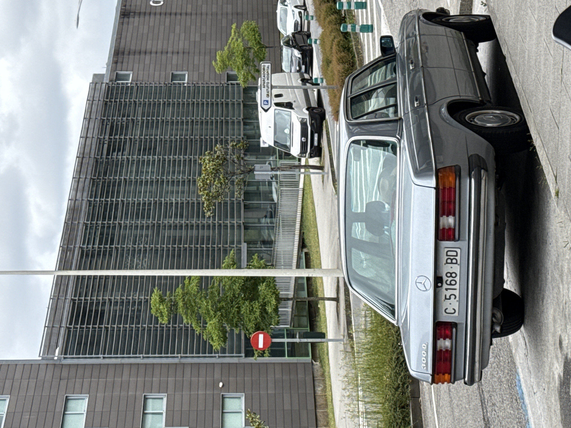The image presents an urban street scene in Santiago de Compostela, Spain, under an overcast sky. In the foreground, a silver Mercedes-Benz 300 D four-door sedan is parked diagonally on the right side of the asphalt road, with its rear wheels partially on the concrete sidewalk. The license plate "C 5168 BD" is clearly visible on the rear of the vehicle.
In the midground, a white utility pole stands centrally. To its left, a red circular "No Entry" traffic sign with a white horizontal bar is mounted on a pole, positioned near a small tree. Further down the street on the right, a white Volkswagen van is parked, followed by a black hatchback and a white sedan. A white rectangular sign, indicating "APARACADOIRO GRATUITO HOSPITAL" (Galician for "FREE HOSPITAL PARKING LOT"), points to the right. The roadside features patches of green grass, small bushes, and several trees with green foliage. Green and white striped bollards line the edge of the road near the parked vehicles.
The background is dominated by a large, modern building complex. The central section of the building features a multi-story facade composed of numerous horizontal louvers or shading elements over dark windows. To the left and right of this section, the building exhibits a grey, block-textured facade with rectangular windows. A pedestrian ramp with white railings leads up to an entrance area on the left side of the main building. No individuals are visible within the depicted scene.