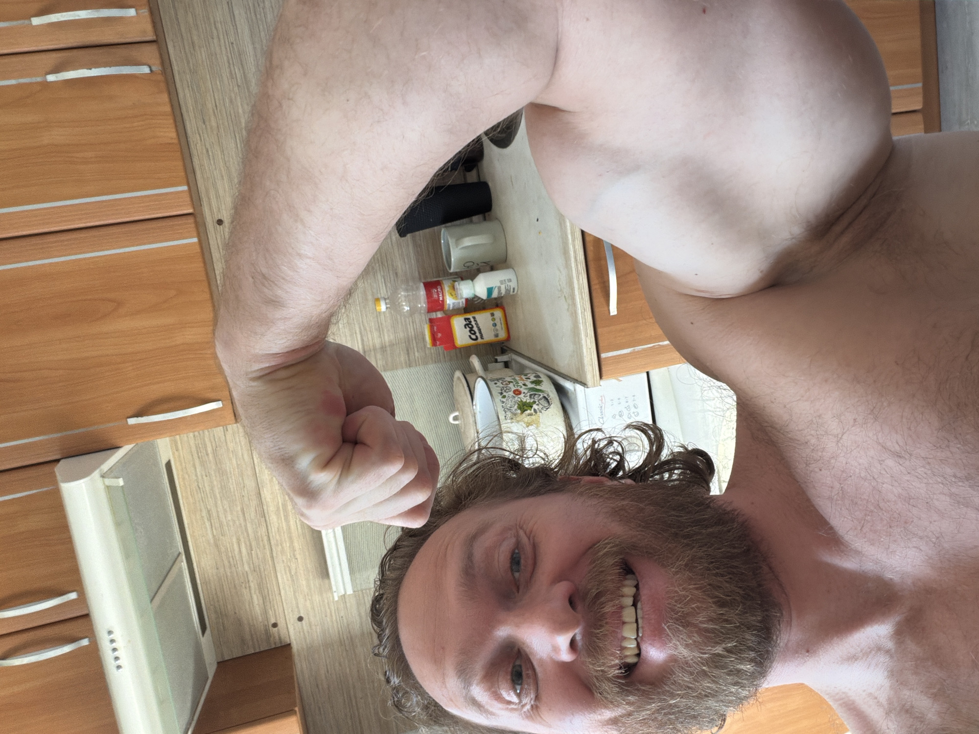A shirtless man with a beard and long hair is flexing his bicep in a selfie. He is standing in a kitchen, evidenced by the presence of wooden cabinets, a range hood, and various kitchen items on the countertop. The kitchen items include bottles and containers. The location cannot be determined as Saint Petersburg based on the provided image.
