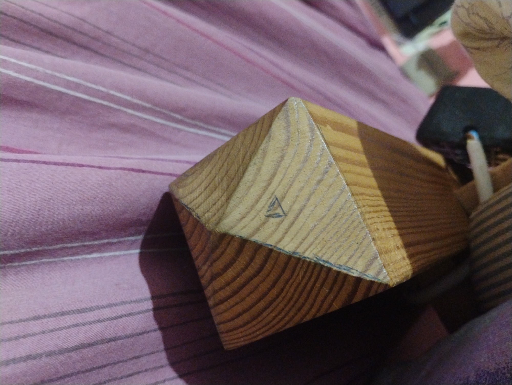 A close-up, top-down view captures the corner of a wooden object, possibly a piece of furniture, positioned against a backdrop of pink fabric with vertical white and grey pinstripes. The wood grain is clearly visible, showing a light brown hue with darker lines. A small, hand-drawn triangle with a line through it is visible on the upper surface of the wood. A shadow is cast from the object onto the fabric. The background fabric appears to be a curtain or upholstery. The image does not contain information about the city of Plachkovtsi, Bulgaria.