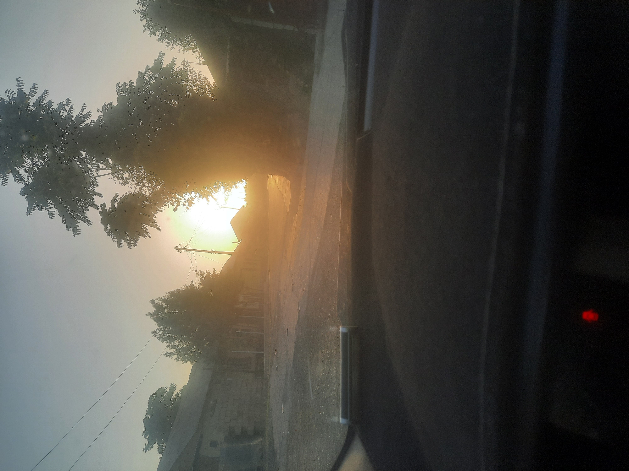 The image captures a street scene at sunrise or sunset in the city of Vosēābād, Tajikistan. The sun is low on the horizon, casting a warm, golden light that creates lens flare and silhouettes the trees and buildings. The view is from inside a vehicle, looking out through the windshield. The foreground shows the dark dashboard of the car with a small red indicator light visible. The middle ground displays a paved street lined with low-rise buildings, some with stone walls, and trees. Power lines are visible in the sky. The atmosphere is hazy, likely due to the low angle of the sun and possibly dust or mist. The environment appears to be a residential or rural area.