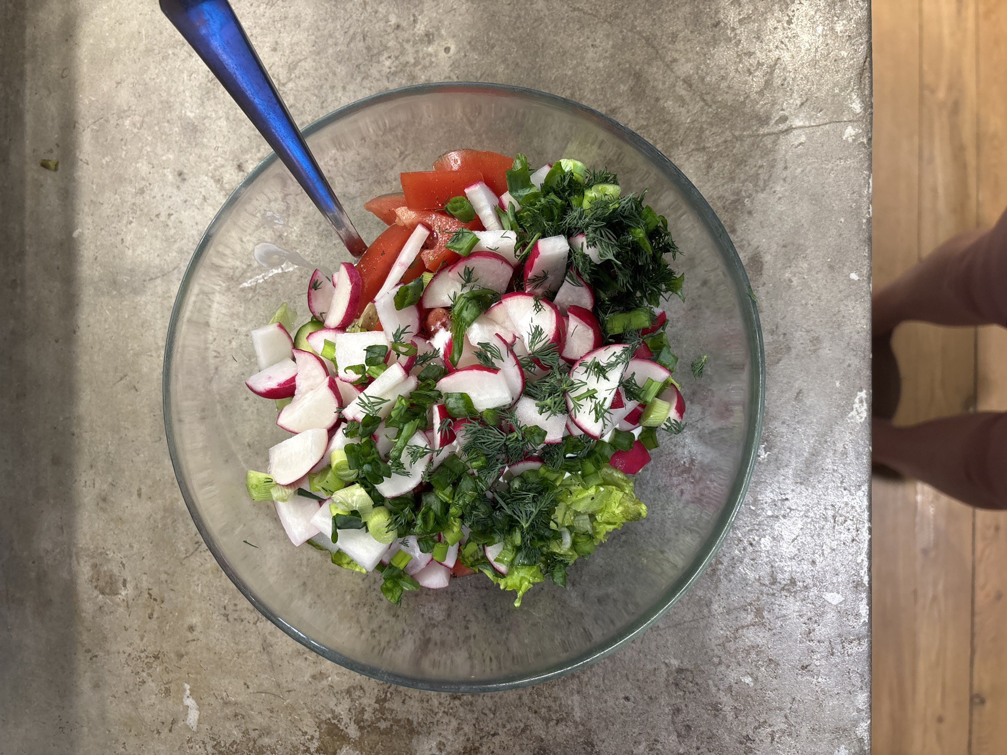 A top-down shot shows a glass bowl filled with a mixed salad. The salad consists of chopped radishes (mostly white with red skin), tomatoes, green onions, and dill. A blue spoon is partially visible in the bowl. The bowl rests on a mottled gray surface. To the right edge of the frame, there is a glimpse of a wooden floor and a person's leg. The location is likely in a kitchen or dining area. The image does not provide specific information to confirm it is located in Chaikovsky, Russia.