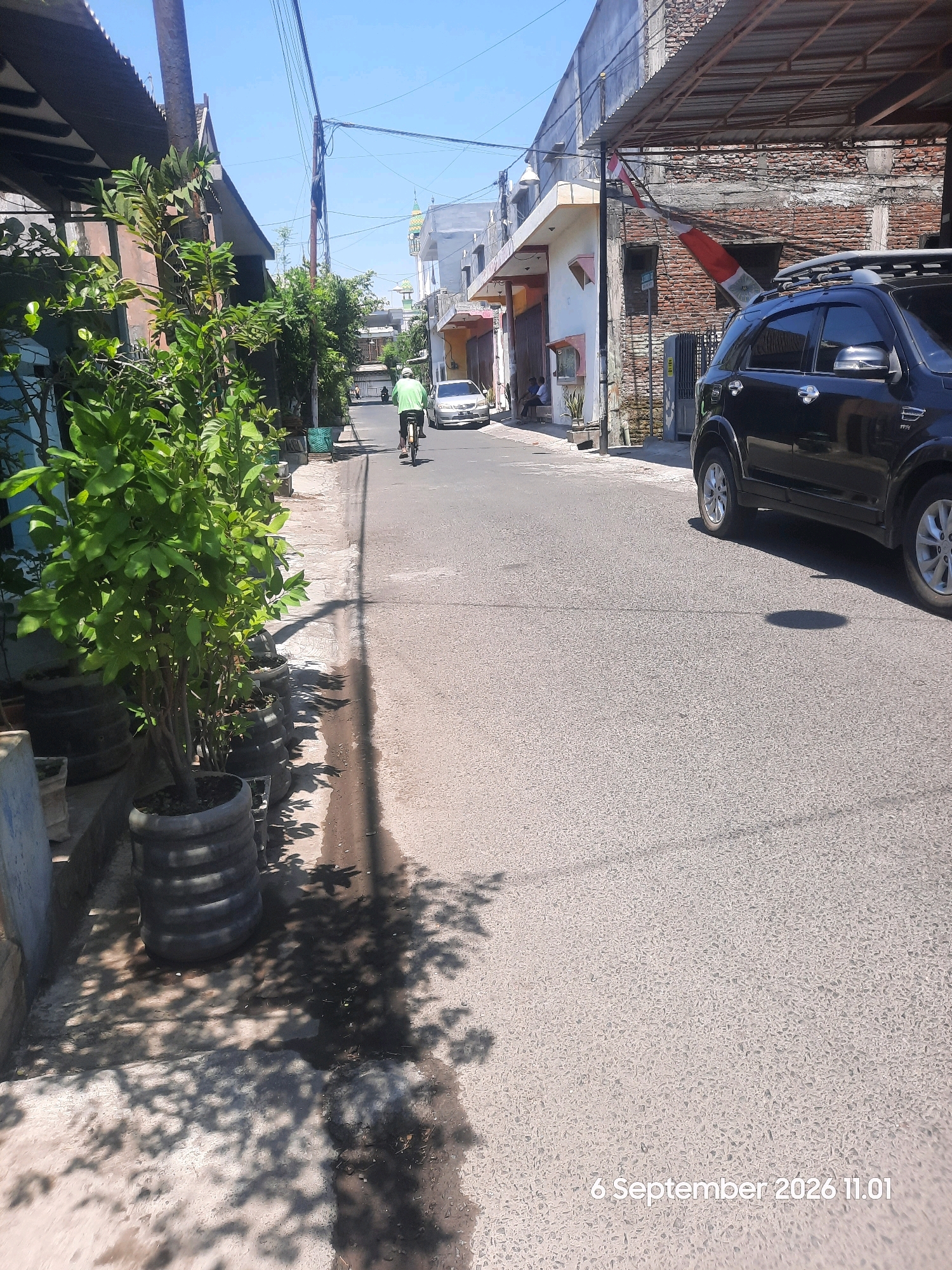 In a sun-drenched street in Probolinggo, Indonesia, a man on a bicycle pedals away from the viewer, his green shirt a bright spot against the asphalt. The street is lined with buildings of varying architectural styles, some appearing to be homes or small businesses. A black SUV is parked on the right side of the road, its metallic sheen reflecting the sunlight. Lush green plants in large pots are positioned along the left side of the street, adding a touch of nature to the urban landscape. The sky above is a clear, vibrant blue, indicating a fair weather day. A timestamp at the bottom right corner reads "6 September 2026 11.01".