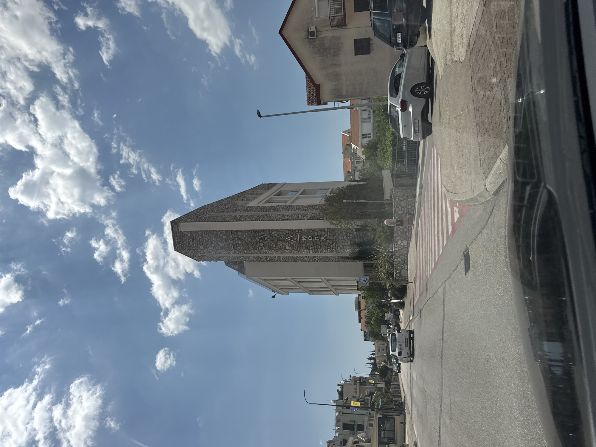 The image captures a street scene in Budva, Montenegro, on a clear, sunny day with scattered clouds. A distinctive building, possibly a hotel or commercial establishment, dominates the right side of the frame. Its facade features a striking combination of light-colored stone and modern architecture. The name "AMORA" is visible vertically on this building.
In the foreground, the perspective is from inside a vehicle, showing the dashboard and windshield. A white Volkswagen Polo is parked near a pedestrian crossing on the right side of the street. A dark SUV is parked behind the Polo. The pedestrian crossing itself is marked with red and white stripes.
The street extends into the distance, lined with other buildings of varying architectural styles and heights. Trees and greenery are interspersed throughout the urban landscape. Several streetlights are visible along the road. A few people are casually walking on the sidewalks. The overall atmosphere suggests a typical day in a coastal town, with a mix of residential and commercial properties.
