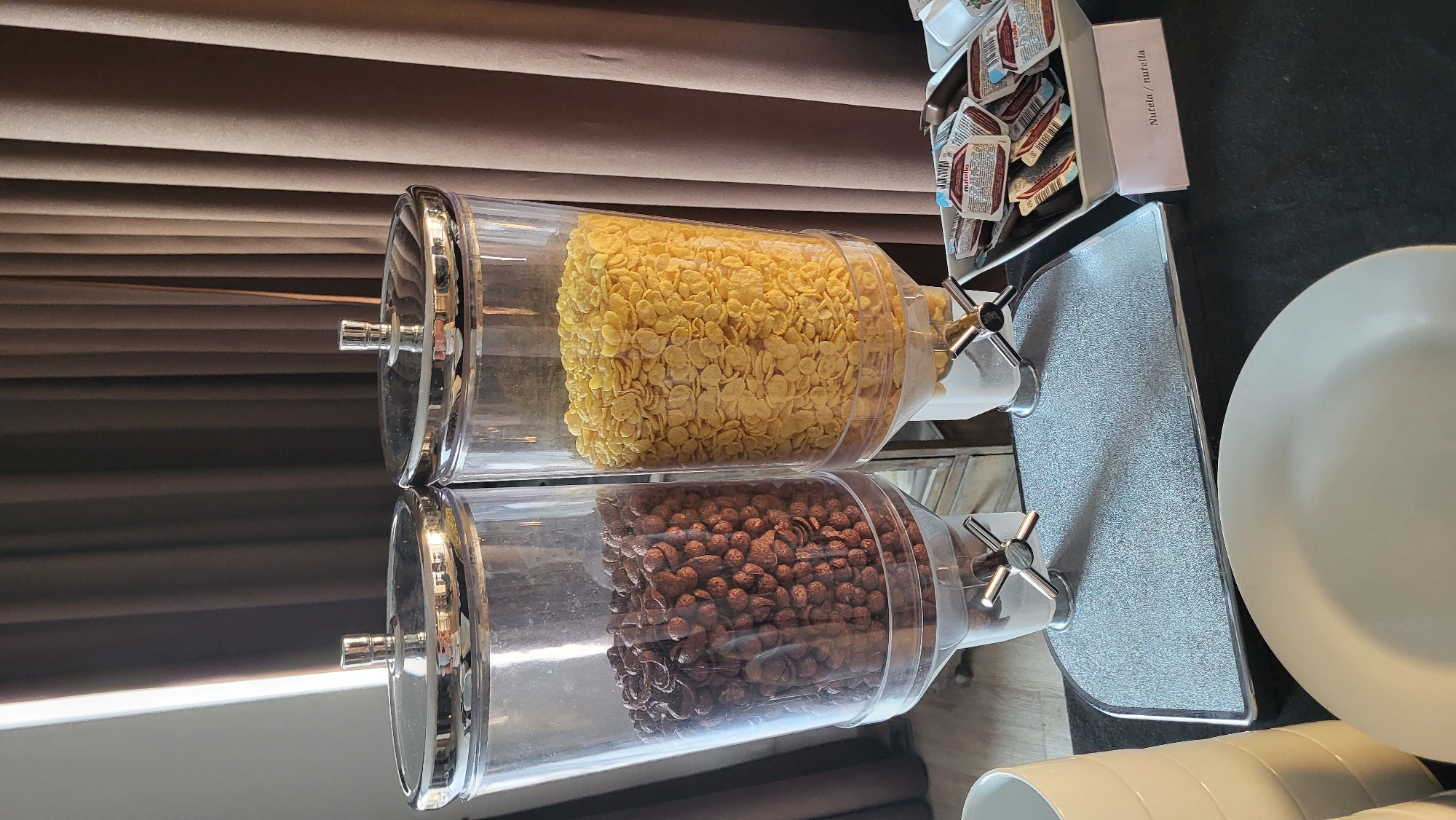 The image shows a breakfast buffet with two large, clear dispensers filled with cereal. The left dispenser contains dark brown, spherical cereal, likely chocolate-flavored puffs. The right dispenser holds light yellow, flake-style cereal, possibly corn flakes. Both dispensers have chrome lids with silver handles and are mounted on a white base with silver dispensing levers.
To the right of the cereal dispensers, there is a white rectangular tray filled with small, individually wrapped portions of Nutella, a hazelnut spread. A small pink label placed in front of the tray reads "Nutela / nutella".
In the foreground, a large, light grey plate is visible, resting on a dark countertop. To the far left, a stack of off-white cylindrical containers, possibly cups or bowls, is partially visible.
The background consists of dark brown, pleated curtains. The lighting suggests an indoor setting, likely a hotel or restaurant.
Based on the provided information that the image is from Prešov, Slovakia, this scene is likely depicting a breakfast service in a hotel, restaurant, or catering facility located in Prešov.
