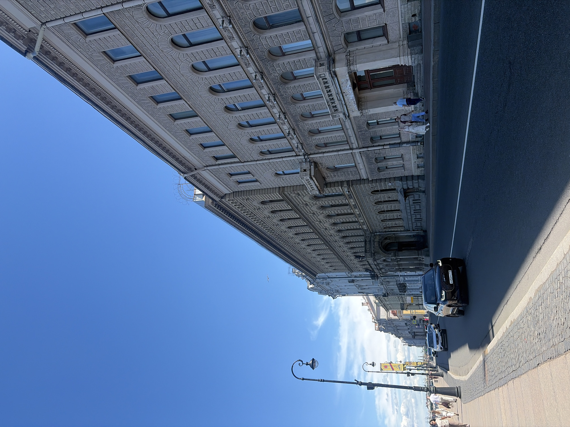 This image captures a sunlit street scene in Saint Petersburg, Russia, observed from a low angle looking down an urban thoroughfare. The clear blue sky dominates the upper portion, featuring a few wispy white clouds and a small bird in flight.
On the right side of the image, a continuous row of multi-story classical-style buildings with ornate facades lines the street. These buildings are constructed from light-colored stone or stucco, featuring decorative cornices, patterned brickwork, and numerous arched windows, many with blue-tinted glass. Several balconies are visible, some with elaborate railings. A prominent arched entrance is seen on one of the buildings further down.
The street, paved with dark asphalt, occupies the central and lower-right portions of the frame, with a visible white dashed lane marking. A black Audi Q8 SUV with Russian license plate `A492PY778` is prominently featured in the middle ground, driving away from the viewer. Behind it, other vehicles, including a white SUV and a dark sedan, are visible further down the street.
On the left, a wide pedestrian sidewalk runs parallel to the street. The section nearest the curb is paved with cobblestones, transitioning to smoother flagstones further back. Several pedestrians are visible on this sidewalk, walking away from the camera. Two distinct black, ornate streetlights with lamps are positioned along the left edge, one in the foreground and another further back. A yellow banner hangs from one of the streetlights.
Near an entrance on the right sidewalk, three individuals are present: a woman in a white dress, a man in a blue shirt and jeans, and another man in a light blue shirt and dark pants. The overall impression is a bustling urban environment on a bright, clear day in a historic city.