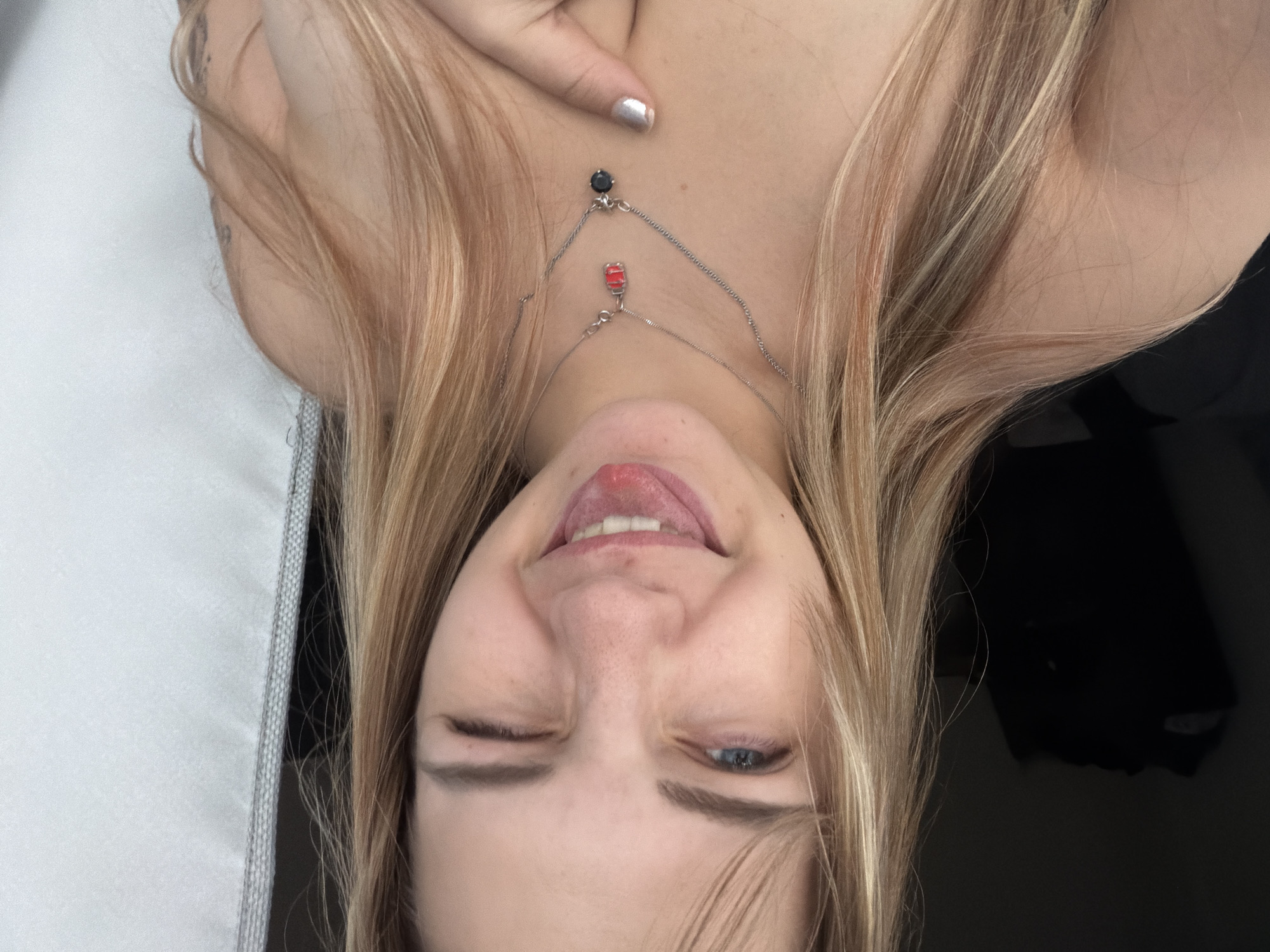 A head-and-shoulders view of a person with light blonde hair and blue eyes. The person is winking with their right eye and sticking out their tongue. They are wearing a necklace with two pendants, one red and one black. A manicured finger with silver nail polish is visible near their upper chest. The background is a combination of white and dark solid areas. The image is taken from an inverted angle. There is no indication of the city of Kazan, Russia.