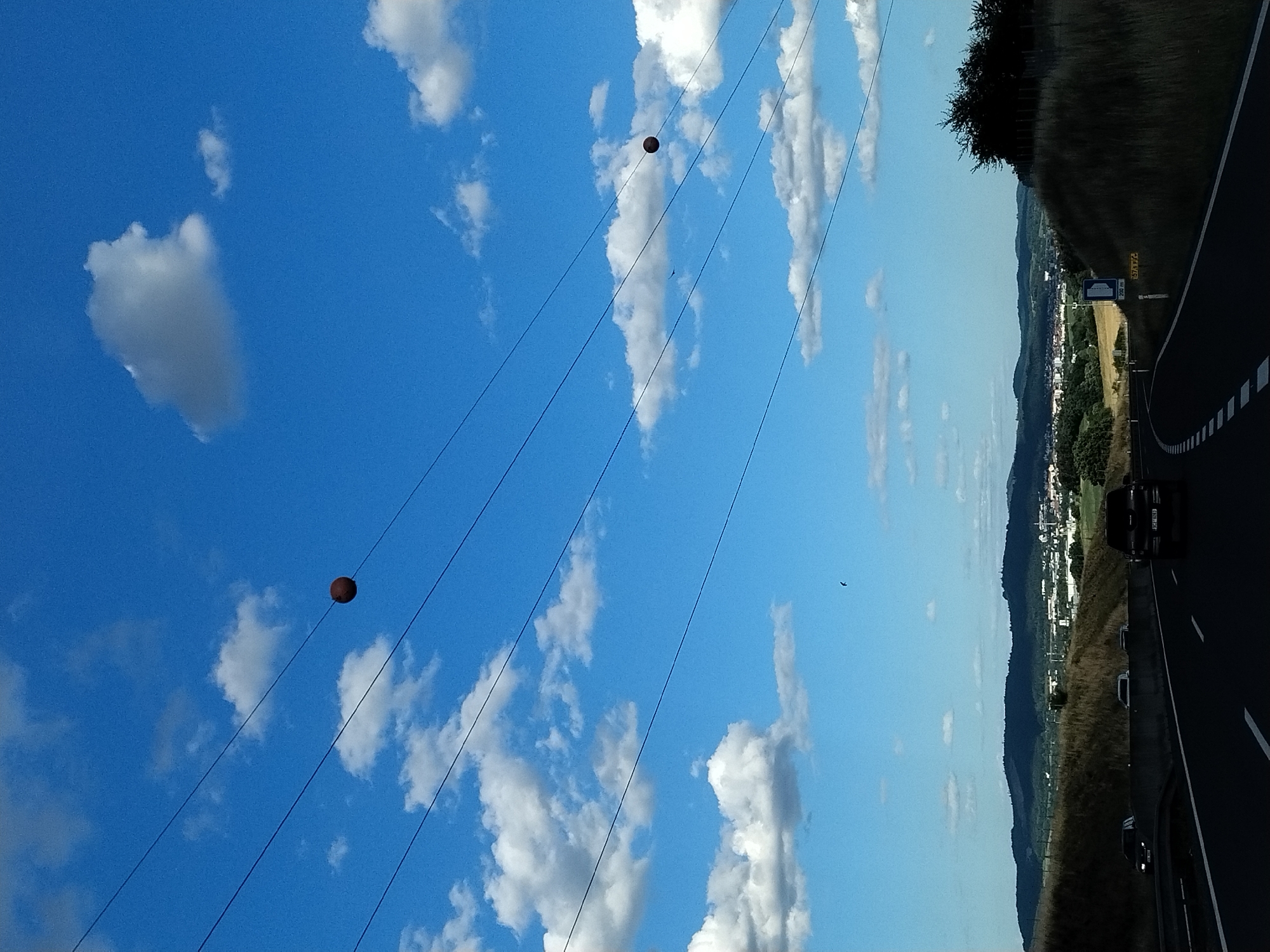 The image shows a bright blue sky with scattered white clouds. Several power lines stretch across the frame. Two dark, spherical objects, possibly weights or markers, hang from the power lines. In the distance, a landscape with rolling hills, fields, and a town is visible under a hazy sky. A highway is in the foreground, with cars driving on it. The scene appears to be a rural or semi-rural area, possibly on the outskirts of Mladotice, Czechia, given the context. The overall impression is one of a sunny day with a highway view.