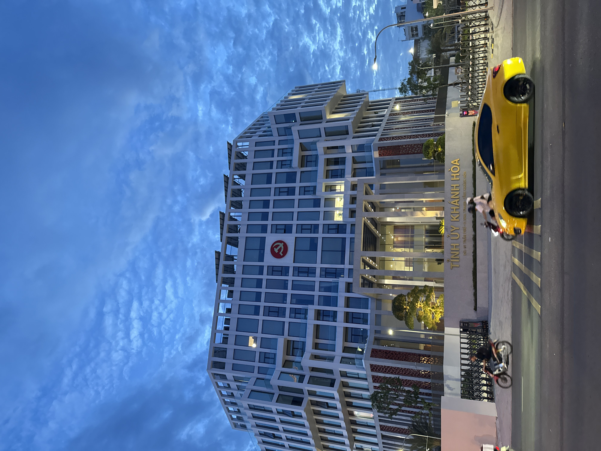 In Nha Trang, Vietnam, at dusk, a striking, modern, multi-story building dominates the skyline, its facade a grid of white panels and large windows, some illuminated. A prominent red hammer and sickle emblem is displayed on the building's upper section. Solar panels are visible on the roof. In front of the building, a low wall with elaborate metal gates spans the property line.
On the street in front of the building, a bright yellow coupe car is in motion, its black wheels blurred from speed. A person is riding a motorcycle to the left of the car, also in motion. Another motorcycle is visible further left, also moving. The street has yellow lane markings. The sky above is a deep blue, filled with textured, scattered clouds that are illuminated by the fading light.