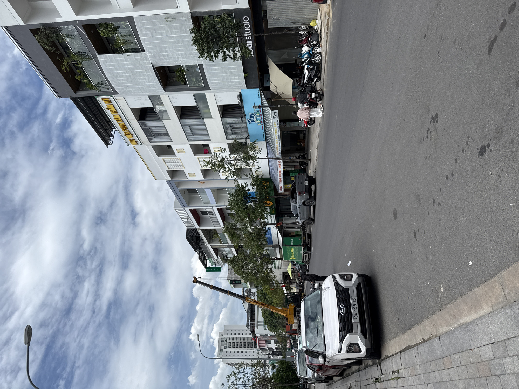 The image captures a street scene in Nha Trang, Vietnam, on a cloudy day. A crane is positioned on the right side of the road, its boom extended upwards. Several vehicles are present, including a white SUV with license plate 78A-195.15 parked on the left, a silver sedan, and a gray pickup truck. A green delivery truck is also visible. On the sidewalk, two individuals are standing near a row of parked motorcycles. The buildings lining the street are modern, multi-story structures with balconies and various businesses on the ground floor, including one with a blue sign that reads "Tany" and another with the text "studio." Trees are planted along the sidewalk. The sky is filled with scattered clouds.