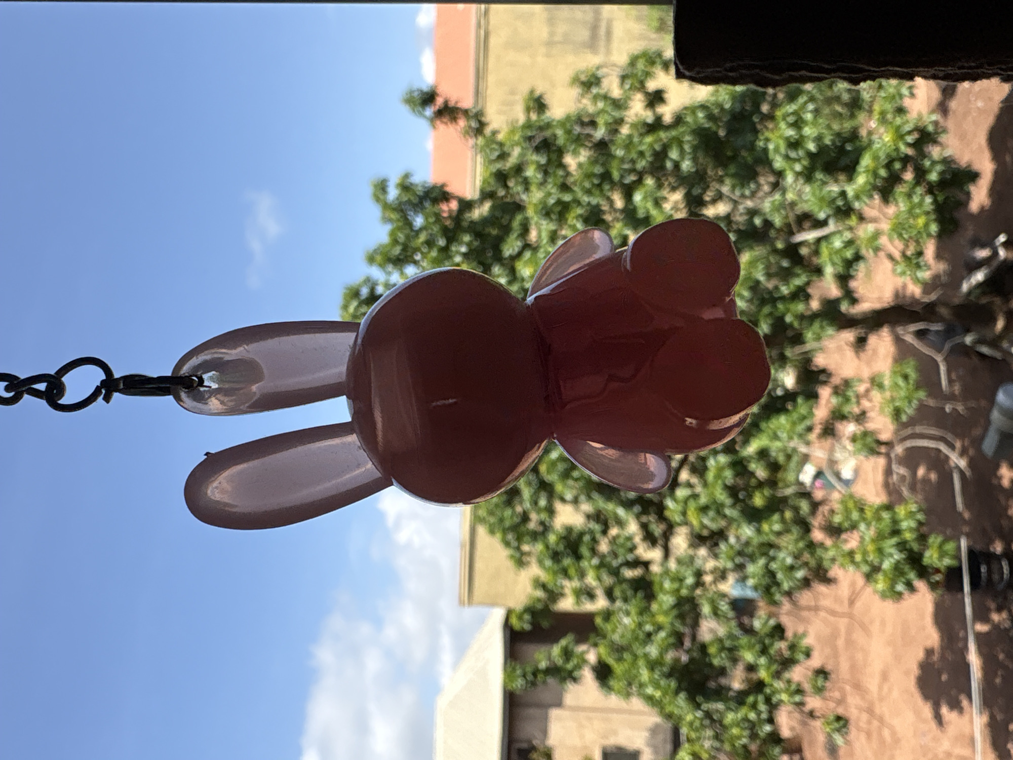 A pink, translucent bunny-shaped ornament is suspended from a black chain. The ornament is oriented vertically, with its ears pointing downwards. The bunny's body appears to be hollow, with visible seams from its mold. Behind the ornament, a clear blue sky with scattered white clouds is visible. In the background, there is a blurred view of lush green trees and buildings. The buildings appear to have beige and orange facades. The overall lighting suggests it is daytime. The scene is likely outdoors, possibly hanging from a window or balcony. There is no visible text in the image.