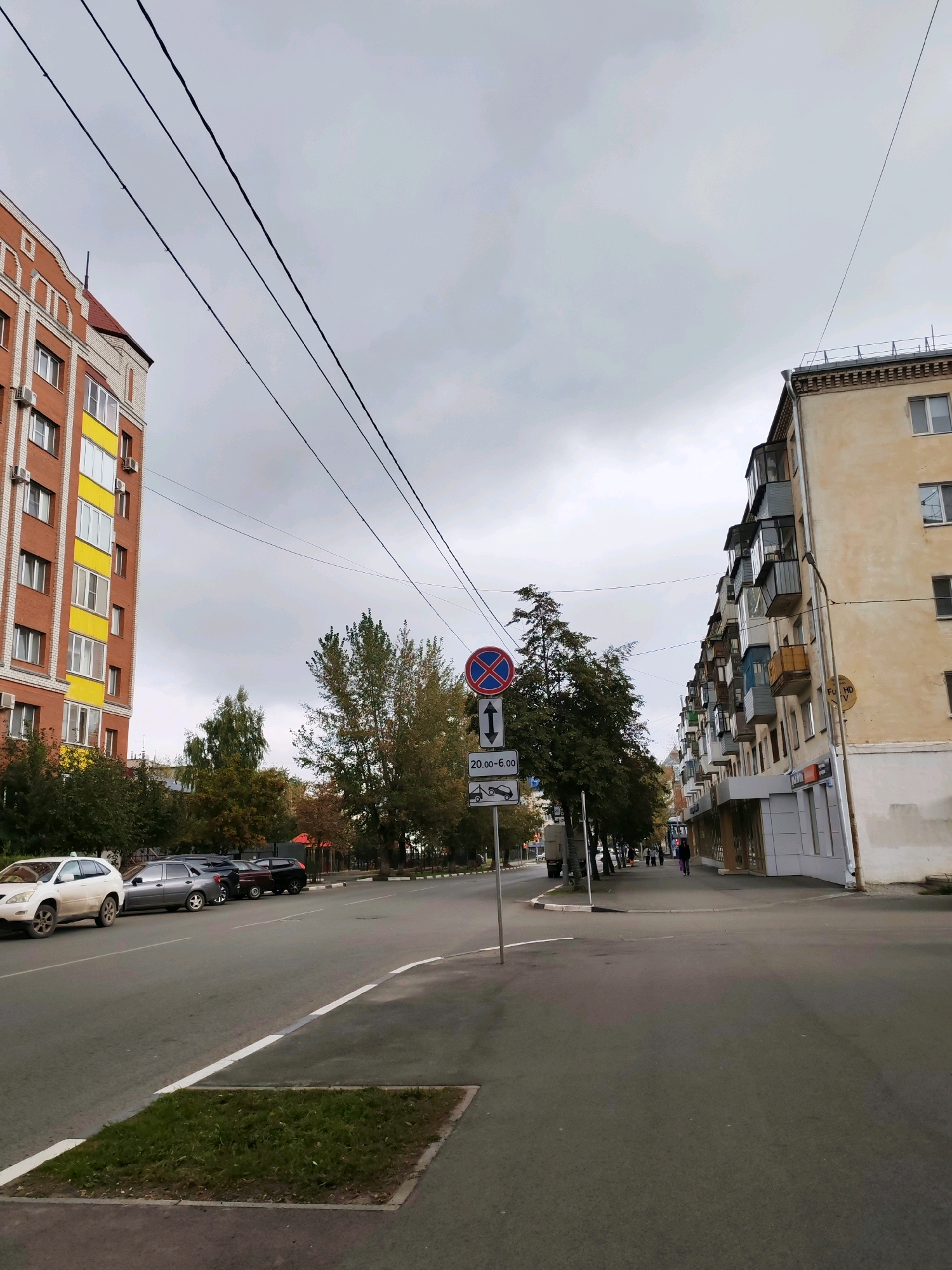 A street scene in a city under an overcast sky. On the left is a multi-story brick building with white trim and yellow accent panels. Across the street on the right is a multi-story building with a pale yellow facade, featuring numerous balconies with colorful railings. A road runs through the center of the image with several cars parked along the right side. A traffic sign with a red circle and a diagonal bar indicating "no stopping" is visible on the left side of the road, with additional signs below indicating time restrictions and towing. Trees with green and yellow foliage line the sidewalk. Power lines stretch across the upper portion of the image.