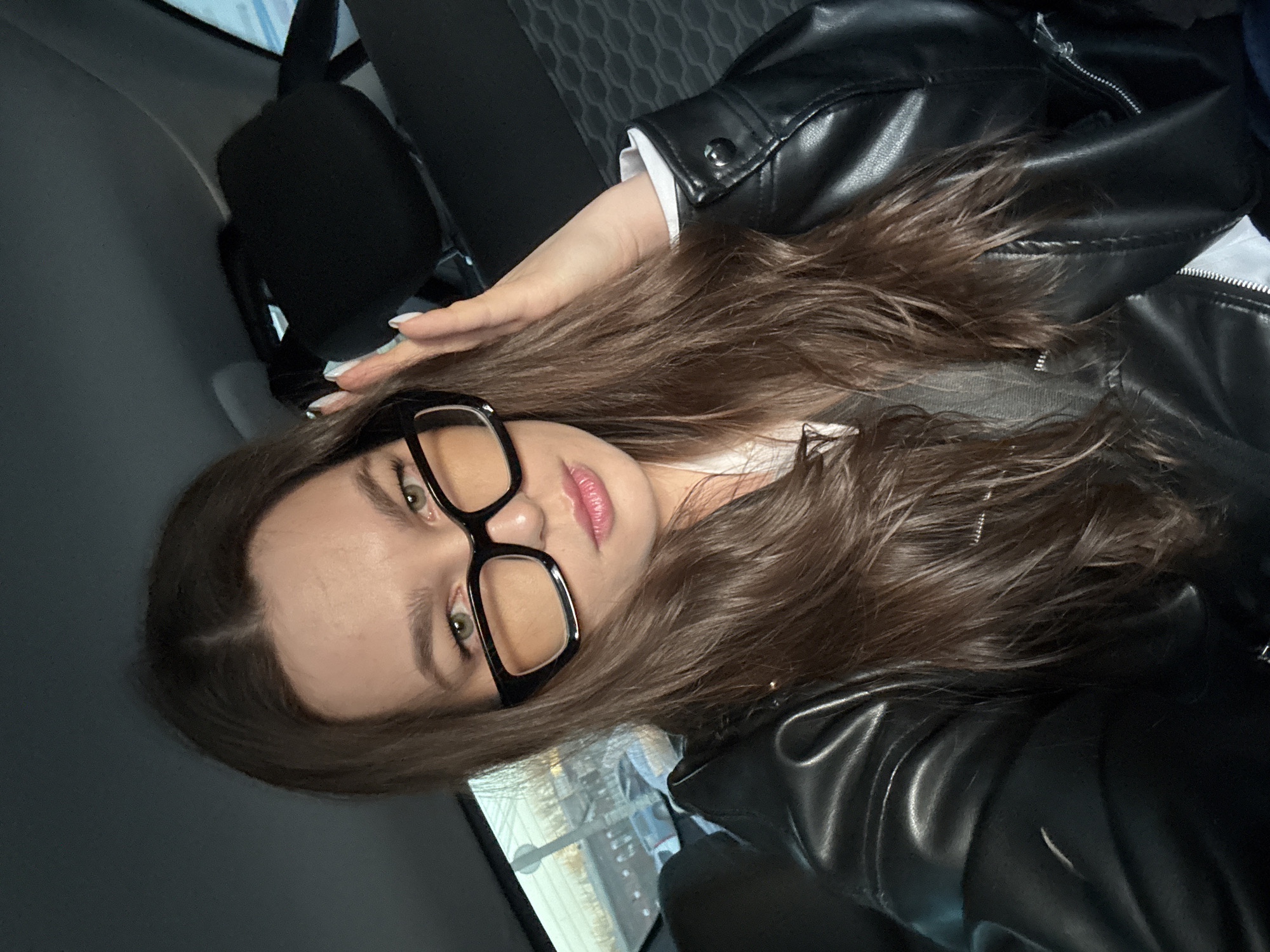 A close-up, angled portrait features a young female inside a vehicle, possibly a car. She has long, wavy dark brown hair and light green eyes. She is wearing large, black-framed glasses and light pink lipstick. Her left hand, with white nail polish, is gently resting on her head. She is dressed in a black leather jacket over a white shirt, with a white cuff visible. The immediate background shows the dark interior of the vehicle, including what appears to be a seat or headrest. A reflection in a window, partially visible behind her, displays urban elements such as lights and structures, suggesting the photo was taken during the evening or night. The individual is looking directly at the camera, posing for the shot. No text is visible in the image. The location context for this image is Izhevsk, Russia.