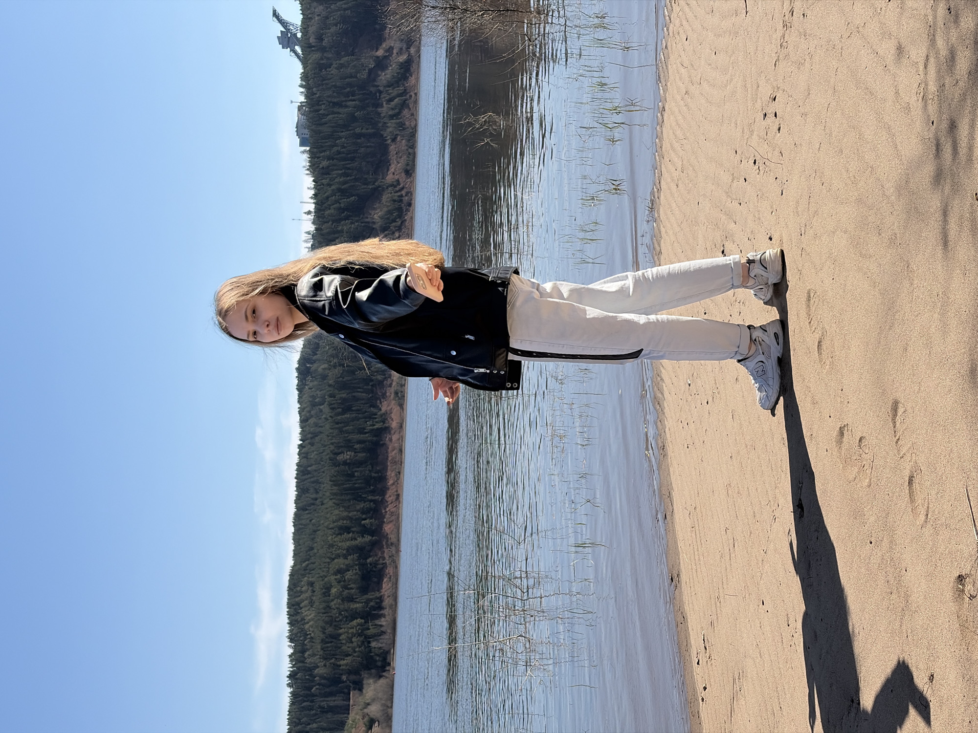 A young woman stands on a sandy beach next to a body of water, potentially a river or lake. She wears a black leather jacket, light-colored pants with a dark stripe, and white sneakers. The background features a forest and a clear blue sky. A crane structure can be seen in the distance, suggesting an industrial presence. The woman is looking at the camera. The location appears to be the city of Noviy, Russia, based on the landscape features and the architecture. The photo was likely taken during a sunny day.