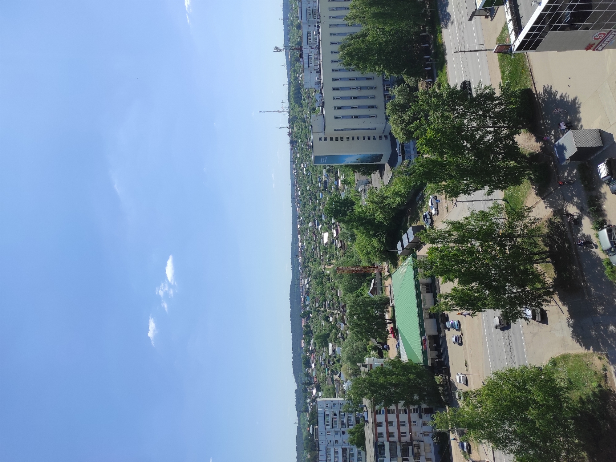 The image depicts a cityscape in Chaikovsky, Russia, taken from an elevated perspective. The sky is blue with scattered clouds. The main urban features include a mix of residential buildings, green spaces comprised of trees and garden plots, and roadways. A prominent building with a distinctive green roof is visible along with a number of cars parked nearby. In the upper part of the image, higher buildings are visible. Infrastructure elements such as utility poles are noticeable. The overall atmosphere suggests a sunny day in a suburban or semi-urban setting.