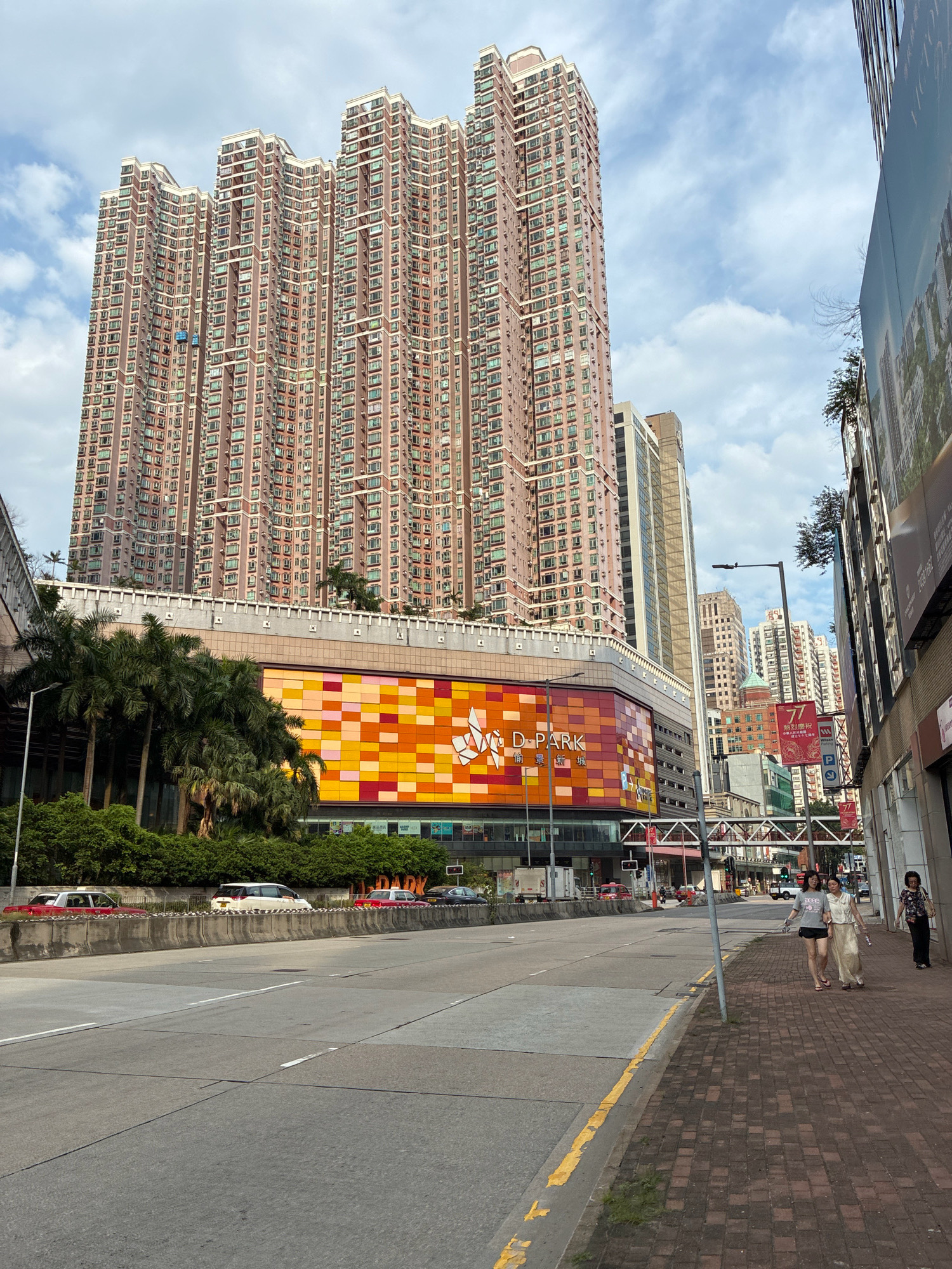 The image depicts a wide street in Hong Kong, China, with a prominent, massive residential skyscraper dominating the background. The skyscraper is characterized by its segmented, wavy roofline and numerous windows. In front of the skyscraper, a colorful, geometric facade of a commercial building, identified as "D-PARK," is visible. Palm trees and lush greenery are situated between the street and the commercial building. On the right side of the street, there is a sidewalk paved with brick-like stones, and two women are walking on it. The street itself is multi-lane with traffic and some parked vehicles. The sky is partly cloudy.
