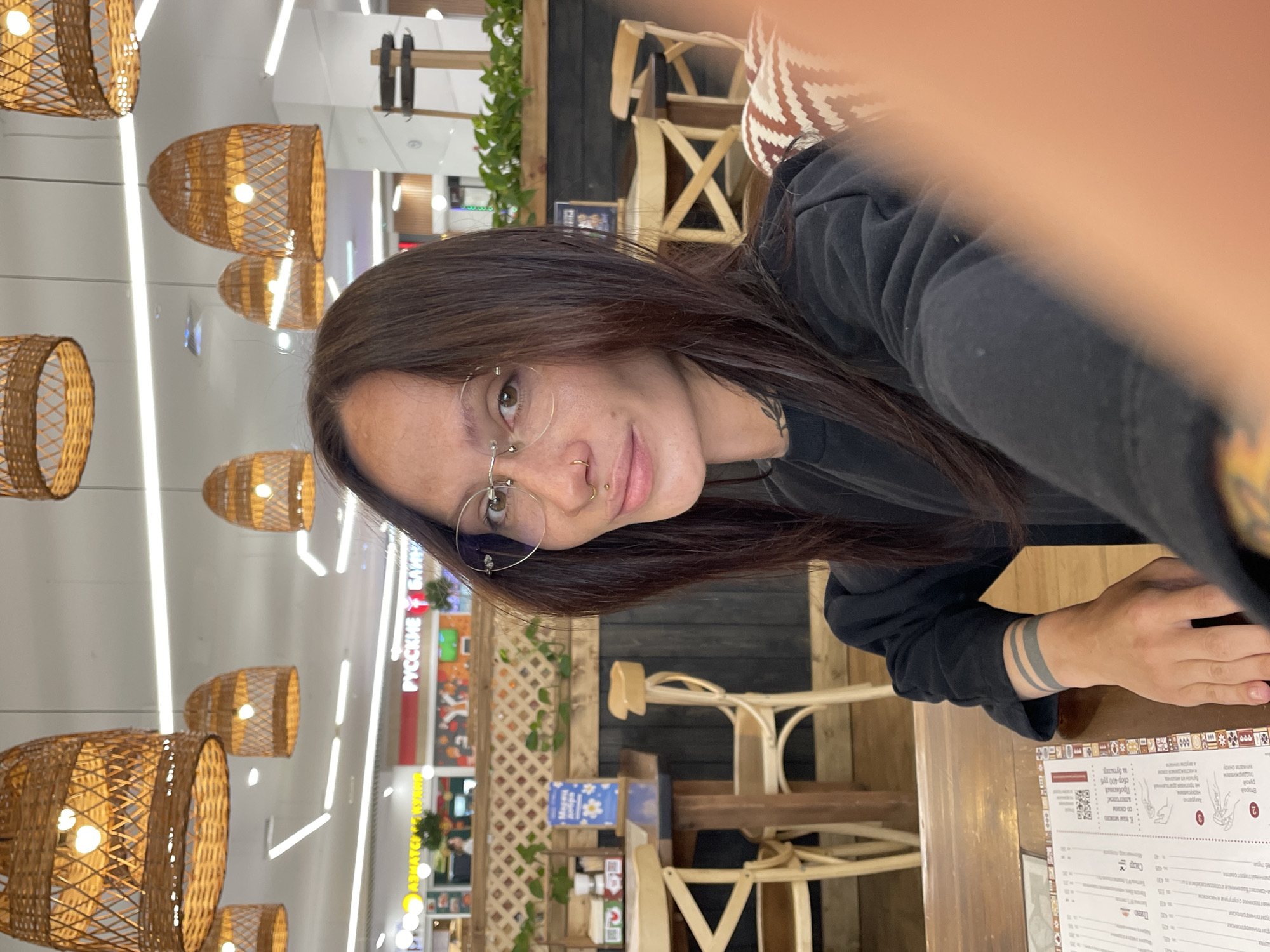 The image depicts a woman taking a selfie in what appears to be a food court or restaurant within a shopping mall in Yekaterinburg, Russia. The woman, positioned centrally, has long, dark brown hair, wears rimless round glasses, a septum piercing, and an upper lip piercing. She is dressed in a black long-sleeved top, with a tattoo of three horizontal gray bands visible on her right wrist and a partial tattoo on her neck. Her right hand rests on a dark wooden table, next to a menu with Cyrillic text. A blurred orange element in the bottom right corner suggests a finger partially obstructing the camera lens.
The background reveals a restaurant setting with light wood chairs, dark wooden tables, and a partition made of wooden planks and a white lattice panel adorned with artificial green vines. A blue sign with white flowers and Cyrillic text is visible on a shelf. Above, numerous large, woven rattan pendant lights with exposed bulbs illuminate the space. The white ceiling also features linear fluorescent lighting. Beyond this immediate seating area, a brightly lit commercial space with various storefronts is visible. Signs in Cyrillic script are prominent, including "РУССКИЕ БЛИНЫ" (Russian Pancakes) and partially visible "АЗИАТСКАЯ КУХНЯ" (Asian Cuisine), consistent with a mall food court environment. A wooden coat stand or decorative structure is visible to the right. A patterned cushion with brown and white geometric shapes is partially visible to the woman's right, suggesting a padded seating arrangement.