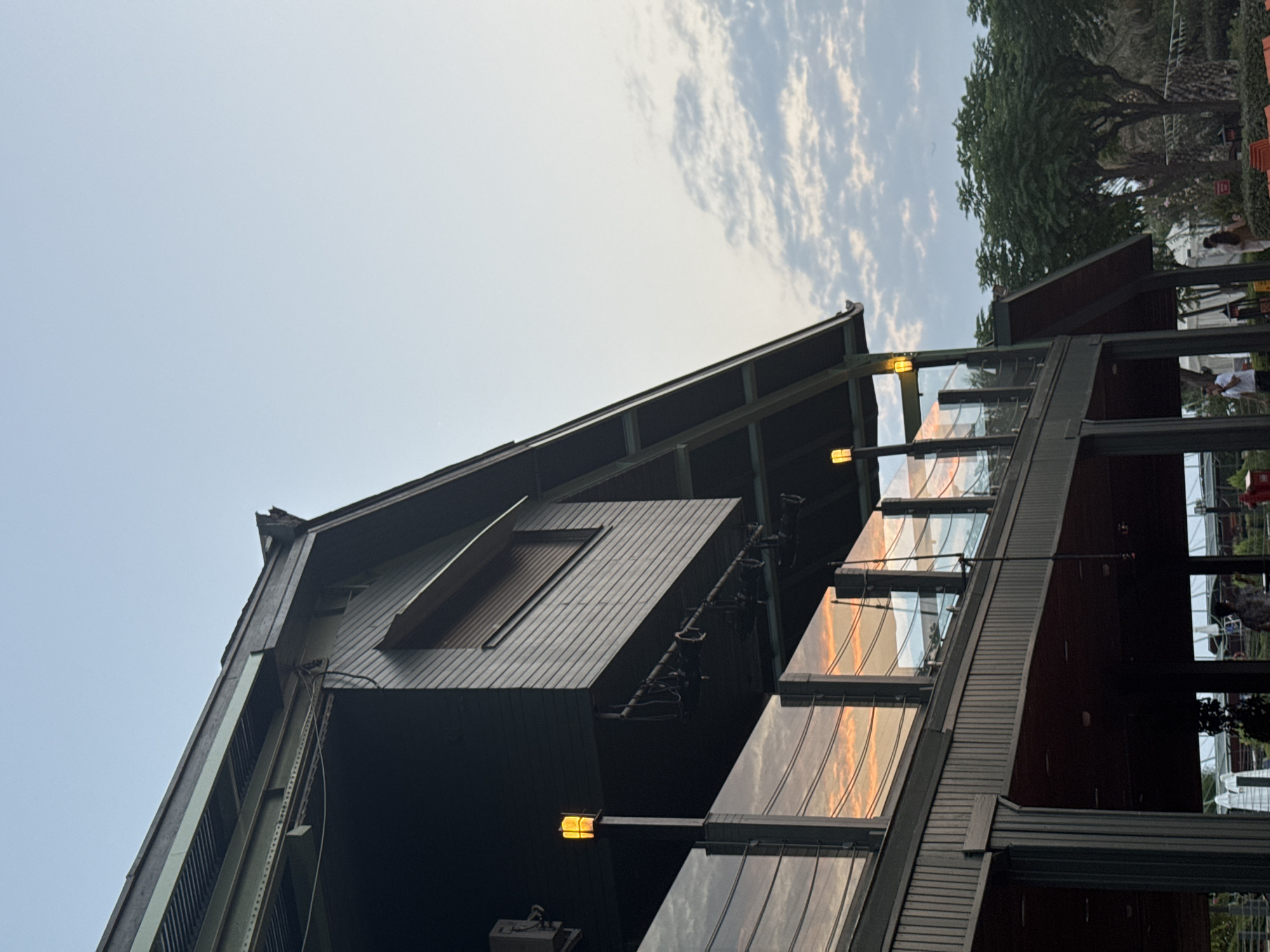 The image shows an outdoor scene with a modern building that features a dark, wooden exterior and large glass windows. The building has a sloped roof with visible structural elements and a second-story balcony or walkway with a glass railing. A warm-toned, square lantern is visible on a pole, illuminating the area. The reflection in the glass windows shows a sky with wispy clouds and the orange hues of a sunset or sunrise. In the background, trees with dense green foliage are present, along with a clear blue sky with scattered clouds. Several people are visible in the lower part of the image, near the base of the building. They are casually dressed and appear to be standing or walking. Some are partially obscured by the building's structure. The overall setting suggests a public space, possibly a restaurant or entertainment venue, in a city that experiences a sunset or sunrise with noticeable color. It is not possible to definitively determine the city of Aksu, Turkey from the visual information alone.