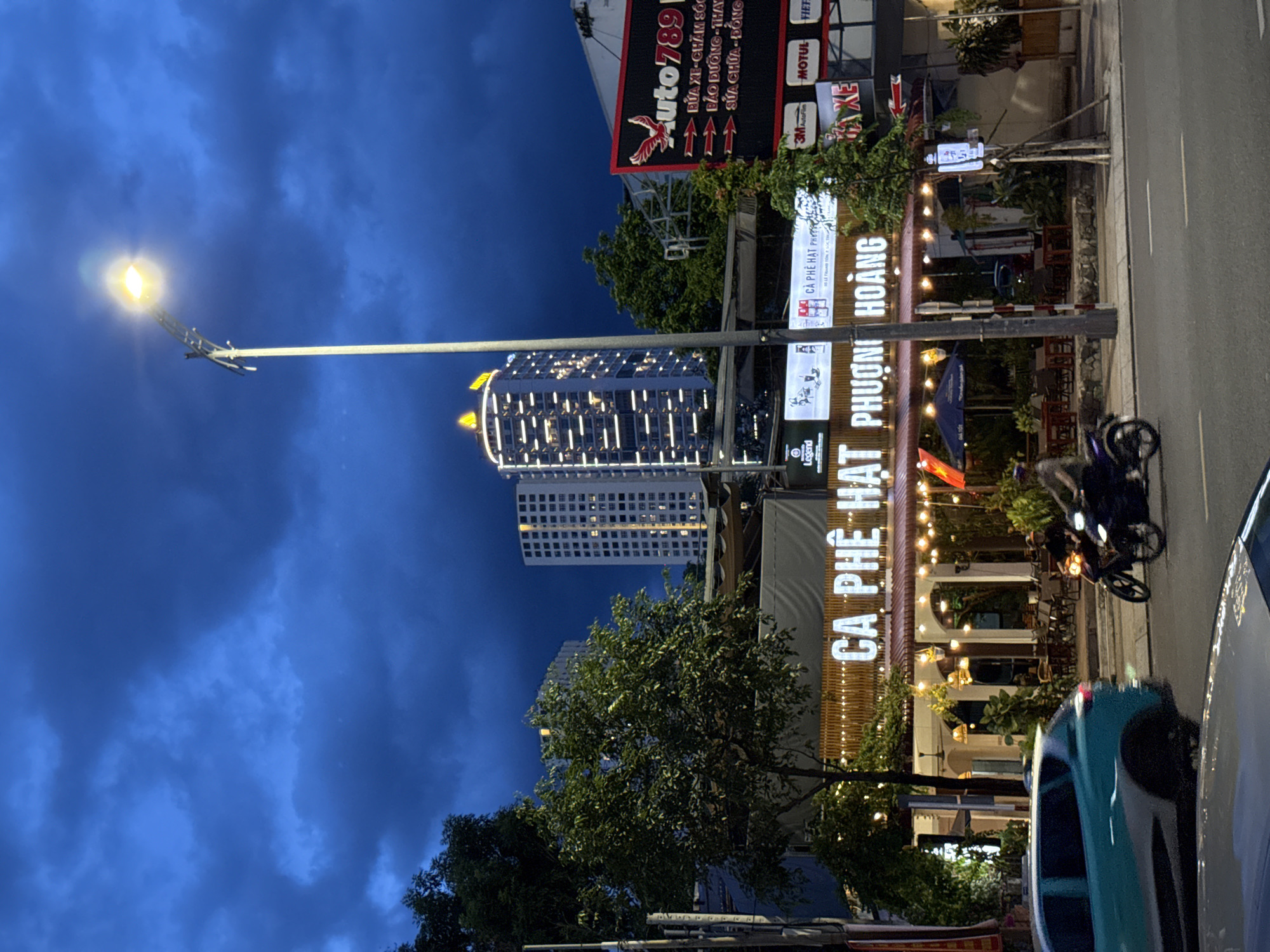 This image captures a street scene in Nha Trang, Vietnam, at dusk. A street lamp with a bright bulb illuminates the dark, cloudy sky. In the background, modern high-rise buildings are lit with numerous windows. In the mid-ground, a building with a warmly lit awning displays illuminated signage that reads "CÀ PHÊ HẠT PHƯƠNG HOÀNG," indicating a coffee shop. The coffee shop appears to have an outdoor seating area with tables and chairs, partially covered by a blue umbrella. A teal-colored car is visible on the left side of the road, and two motorcycles are in motion on the right. One motorcycle has a rider wearing a hat and dark clothing. The overall environment suggests a commercial street in an urban setting, with a transition from daylight to evening.