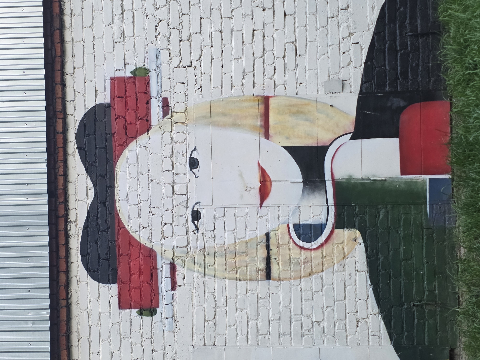 A mural on a white brick wall in Chaikovsky, Russia, depicts a stylized human face with abstract features. The face is white with a round shape, dark eyes, a red triangle for a nose, and yellow and black lines suggesting hair or facial structure. The figure wears what appears to be a black and red hat or head covering and a black suit. The background is primarily white brick, and there is a corrugated metal panel to the left and some green vegetation to the right of the mural. The mural is slightly rotated clockwise in the image.