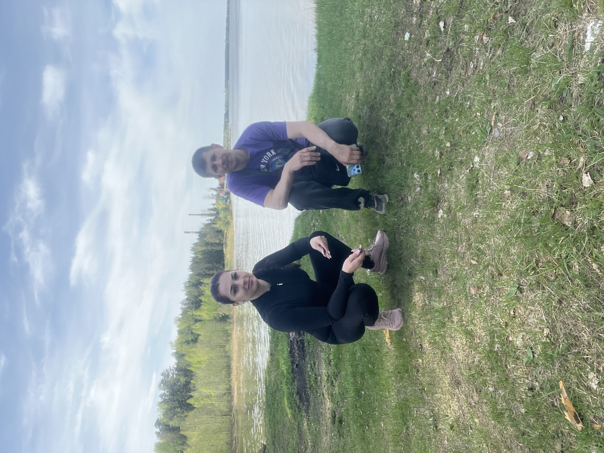 Two individuals, a male and a female, are squatting on a grassy area adjacent to a body of water. The male is wearing a purple T-shirt and dark pants, and holding a smartphone. The female is wearing a black long-sleeved shirt and black leggings, with light brown shoes. The location appears to be near a lake or river, with trees visible in the background. The sky is cloudy. The location may be Chaikovsky, Russia, based on the prompt.