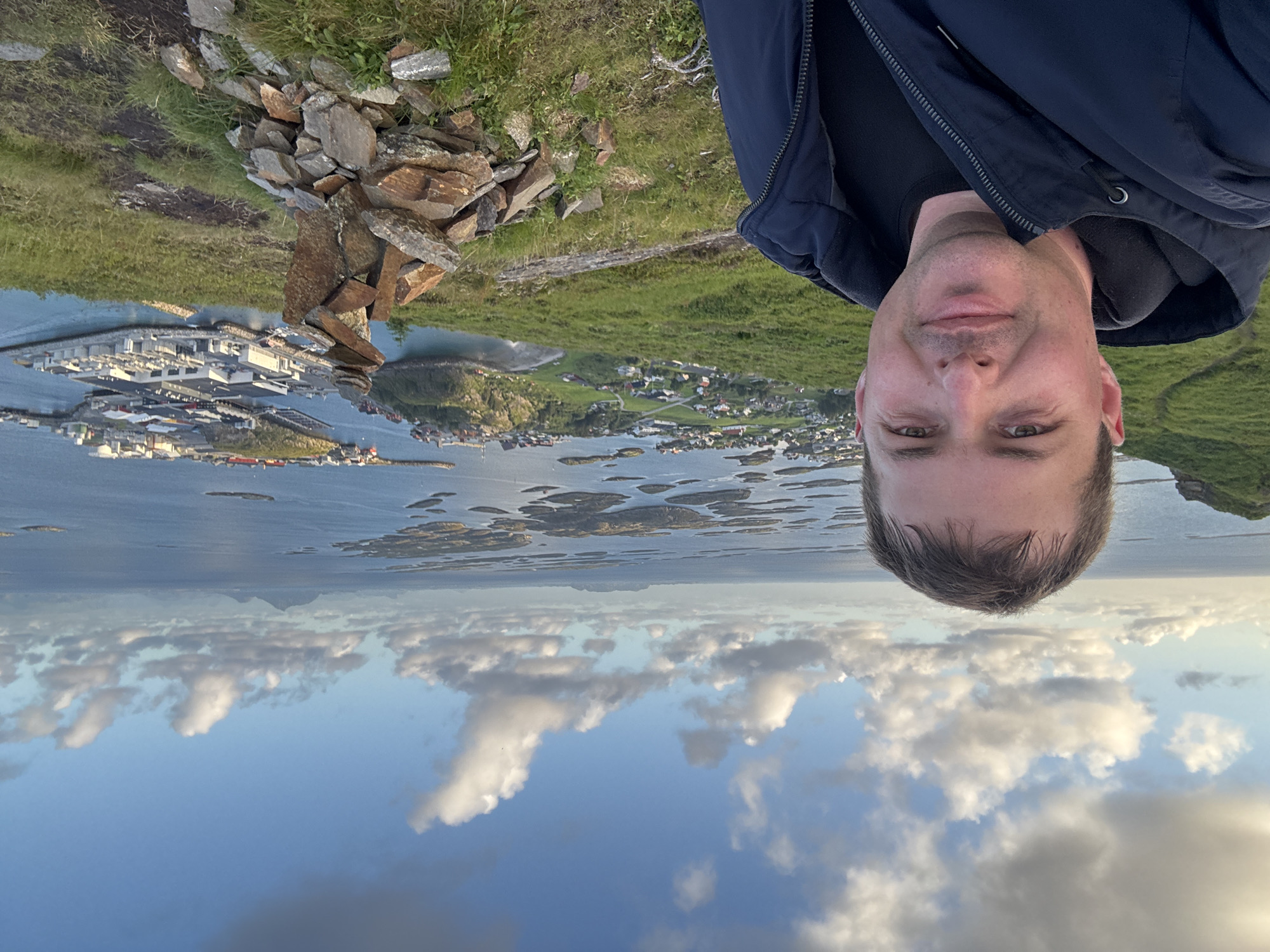 A selfie captured from a high vantage point shows a man in the foreground wearing a dark blue jacket over a black t-shirt. He is looking towards the camera with a neutral expression. Behind him, a panoramic view unfolds of the city of Lovund, Norway, with its characteristic brightly colored houses nestled amongst green hills. The scene extends to the sea, dotted with numerous small islands and skerries. In the distance, larger landmasses rise from the water under a sky filled with scattered white clouds. In the immediate foreground, to the right of the man, a cairn of stones is stacked, suggesting a hiking trail or a viewpoint. The overall environment is coastal and rugged, with a clear blue sky and calm sea.