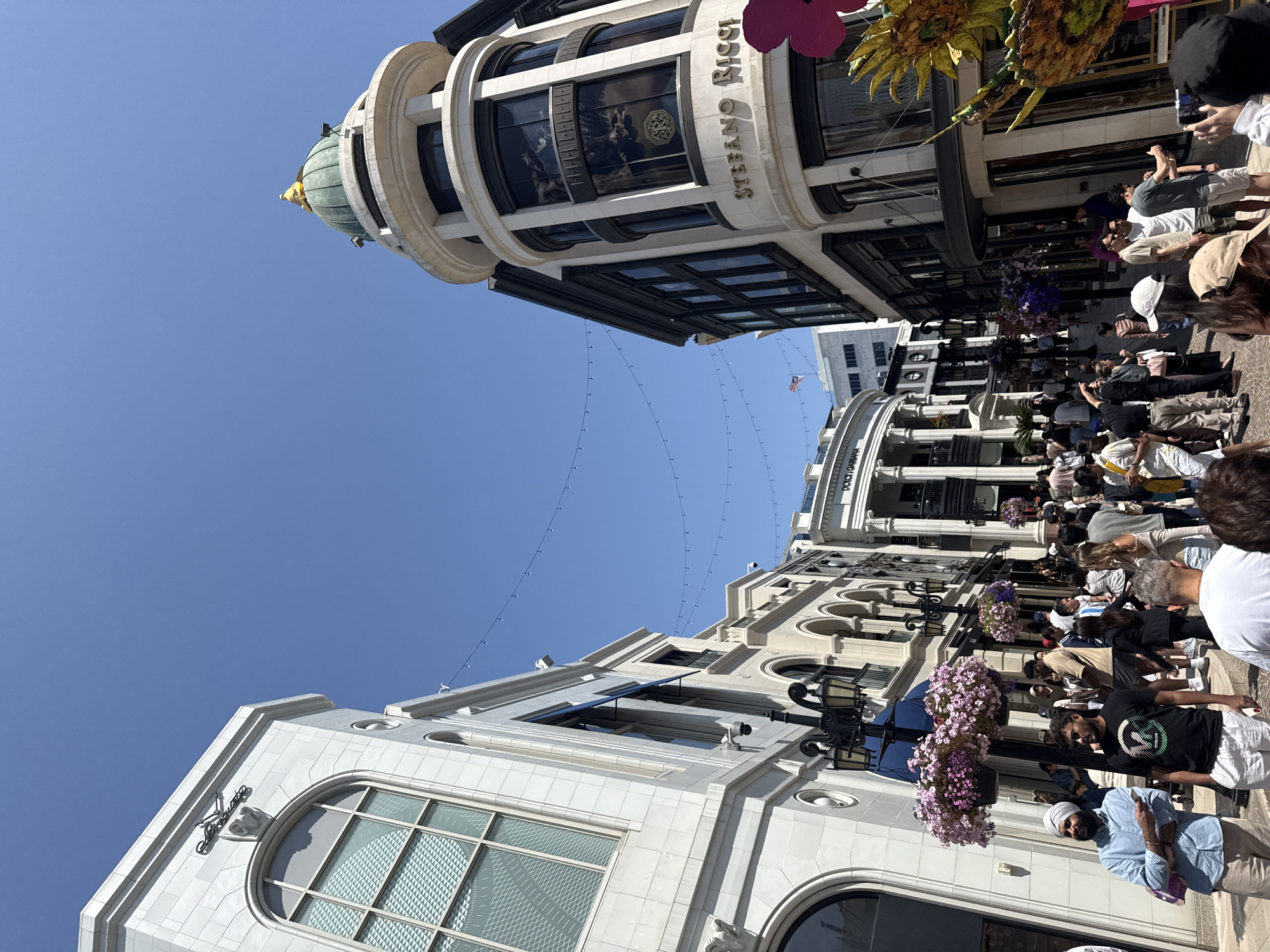The image captures a bustling scene in Beverly Hills, California, United States. The street is lined with high-end retail buildings, predominantly white with ornate architectural details. People are walking along the street, indicating a lively shopping environment. Visible storefronts and building facades suggest a mix of designer boutiques and commercial establishments. Overhead, decorative string lights are strung across the street. The sky is clear blue, suggesting a sunny day. A few decorative planters and flower arrangements add to the visual aesthetic of the area.