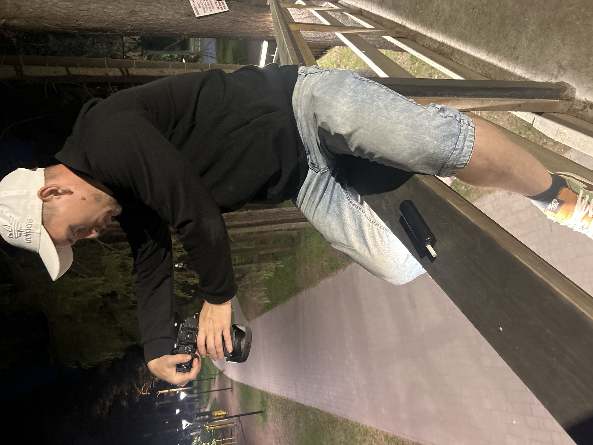 A man is positioned on a wooden railing overlooking a lit walkway in Votkinsk, Russia. He is bent over and holding a camera in his hands, seemingly taking a photograph. He wears a white Adidas cap, a black hooded sweatshirt, acid wash denim shorts, and tan sneakers. A black object is positioned between the man and the railing.