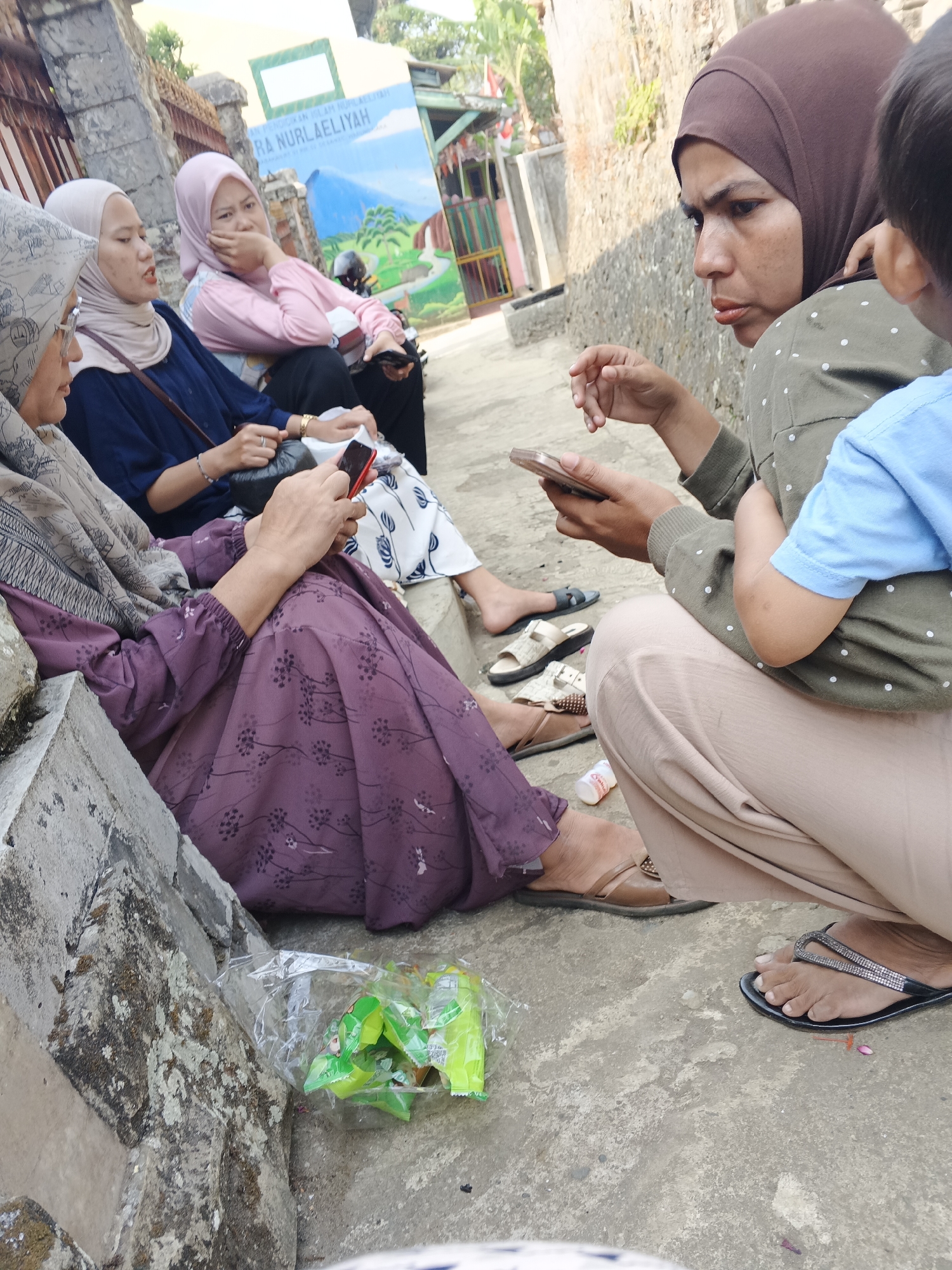 In Warungkiara, Indonesia, three women and a child are gathered outdoors, seated on a concrete surface with a textured stone wall in the background. The woman on the left, wearing a grey hijab and a patterned purple dress, is looking at her phone. Next to her, a woman in a light pink hijab, wearing a dark blue top, rests her chin on her hand, appearing to be in conversation or observation. To the right, a woman with a brown hijab and a green dotted shirt holds a phone and looks intently at it, with a young child in a blue shirt being held by her. Another woman in a light pink hijab is visible behind the woman in the dark blue top. A partially opened plastic bag with green snack wrappers lies on the ground in the foreground. A colorful mural with text in Indonesian is visible on a wall behind the seated individuals. The scene is lit by natural sunlight, casting shadows on the ground.
