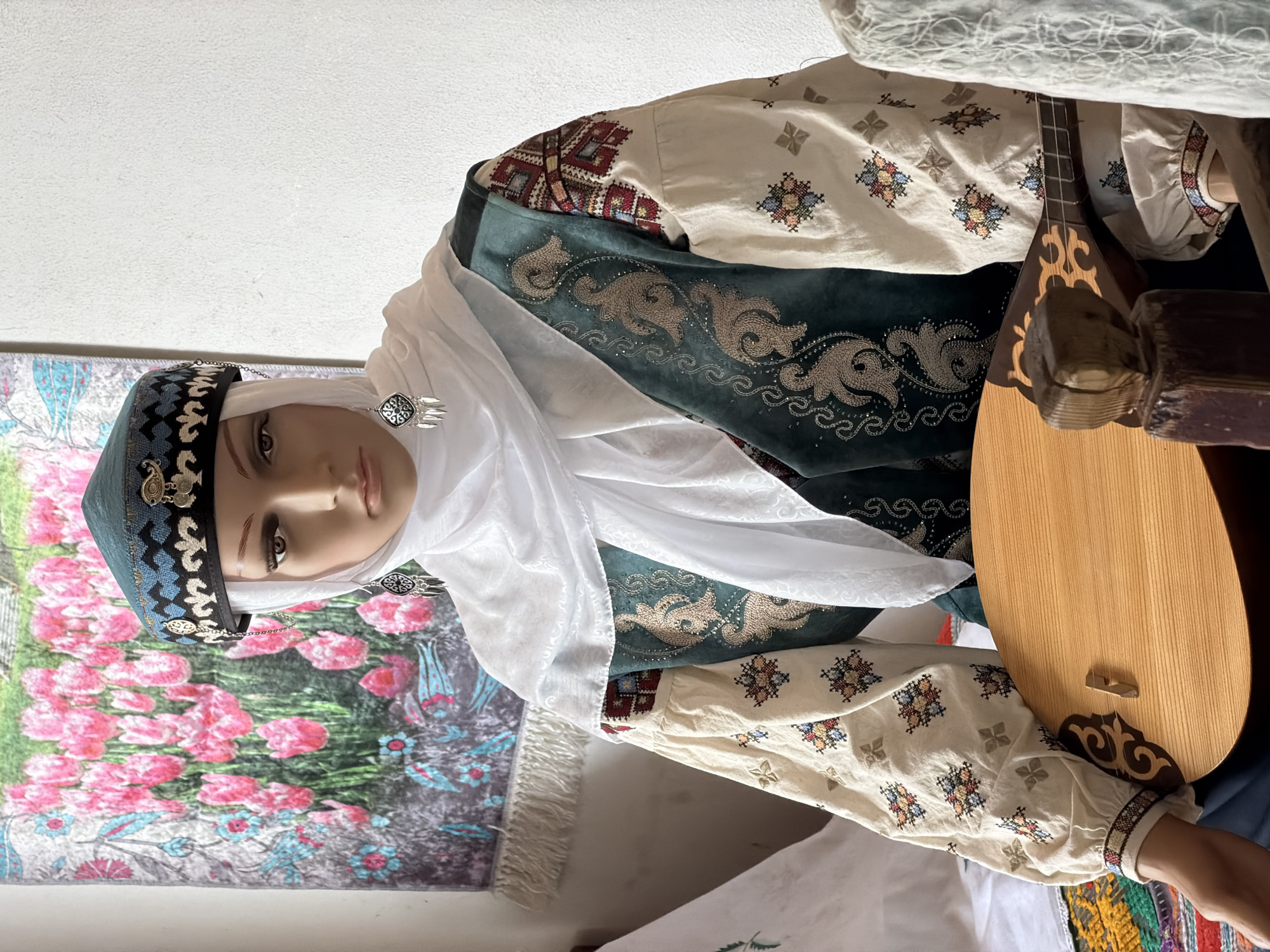 The image shows a mannequin dressed in traditional clothing, likely displayed in a museum or cultural center. The mannequin wears a white hijab and a teal, embroidered cap with a chain detail. The outfit includes a white blouse with embroidered sleeves featuring colorful geometric patterns and a dark teal embroidered vest. The mannequin is holding a light-colored stringed instrument, possibly a lute or a dombra. In the background, a tapestry or rug with a floral pattern, primarily featuring pink tulips, hangs on a white wall. The overall scene suggests a cultural display of traditional attire and music. The location is identified as Літвенскі сельскі Савет, Belarus.