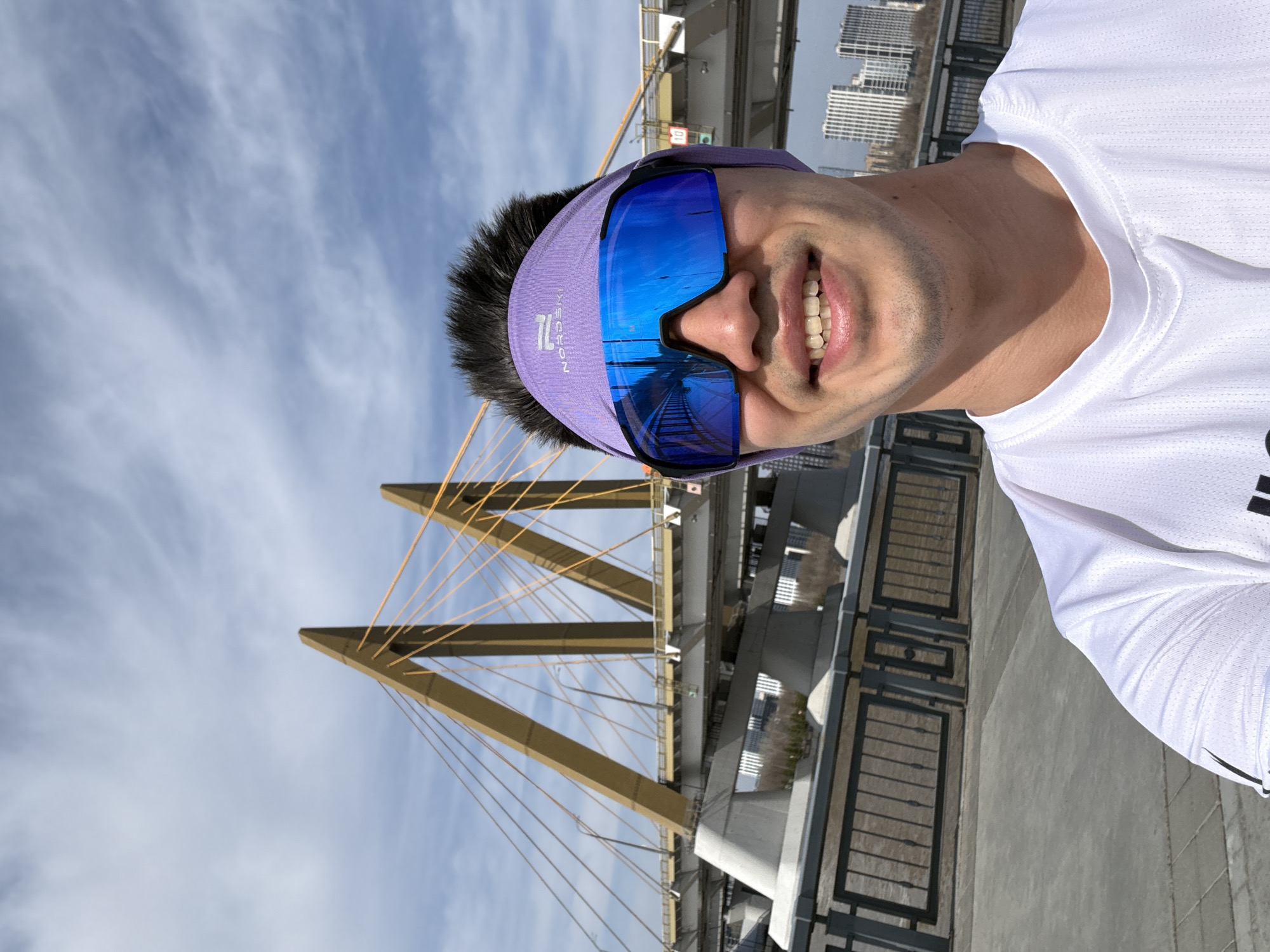 A smiling man in a white athletic shirt and blue sunglasses with a purple headband is the central subject. The sunglasses reflect an image of railway tracks. Behind him is a cable-stayed bridge with yellow support cables. In the background, city buildings can be seen against a blue sky with wispy clouds. The location is in Kazan, Russia, specifically near the Millennium Bridge.