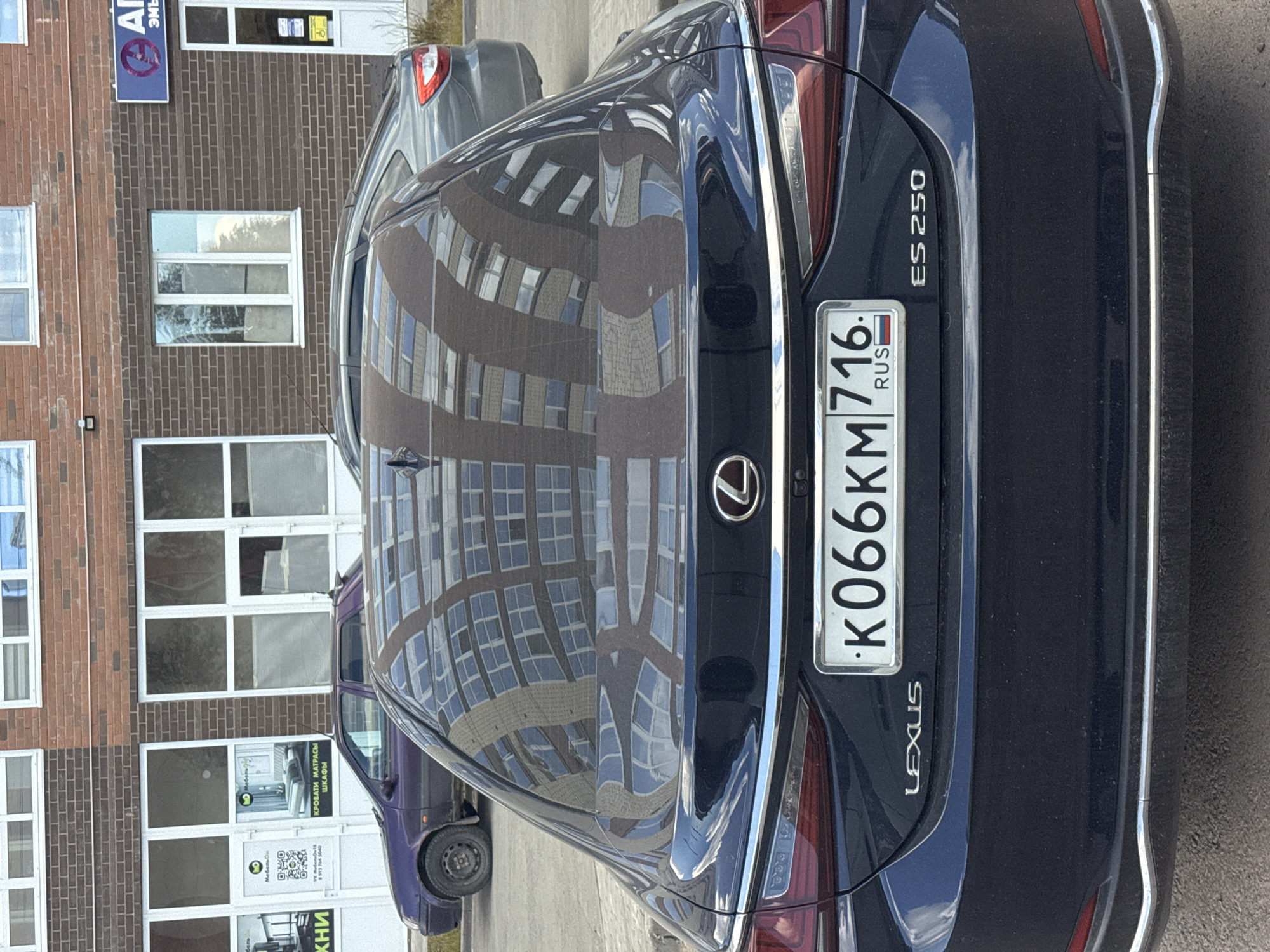 The image depicts the rear of a dark blue Lexus ES 250 parked outside a brick building. The car's license plate reads "K066KM 716 RUS". The building features windows and signs, including "Мебель Он" and "ΑΓ Эмъ". The reflection in the car's paint indicates the presence of another, taller building nearby. A purple minivan is partially visible behind the Lexus. The scene appears to be in Mozhga, Russia, as indicated by the license plate country code.