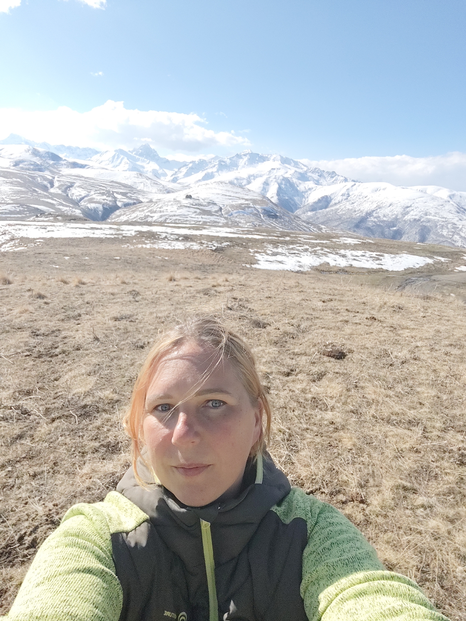 The image shows a selfie taken by a blonde woman wearing a light green sweater with a dark gray vest. The background reveals a mountainous landscape with snow-capped peaks under a clear blue sky with some clouds. The immediate foreground is dry, grassy terrain. Based on the known location, the scene is likely to be in the vicinity of Tyrnyauz, Russia, given the presence of mountains and the time of year suggesting snow cover at higher altitudes.